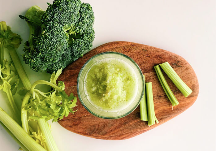A stalk of celery and a head of broccoli next to a glass of celery juice