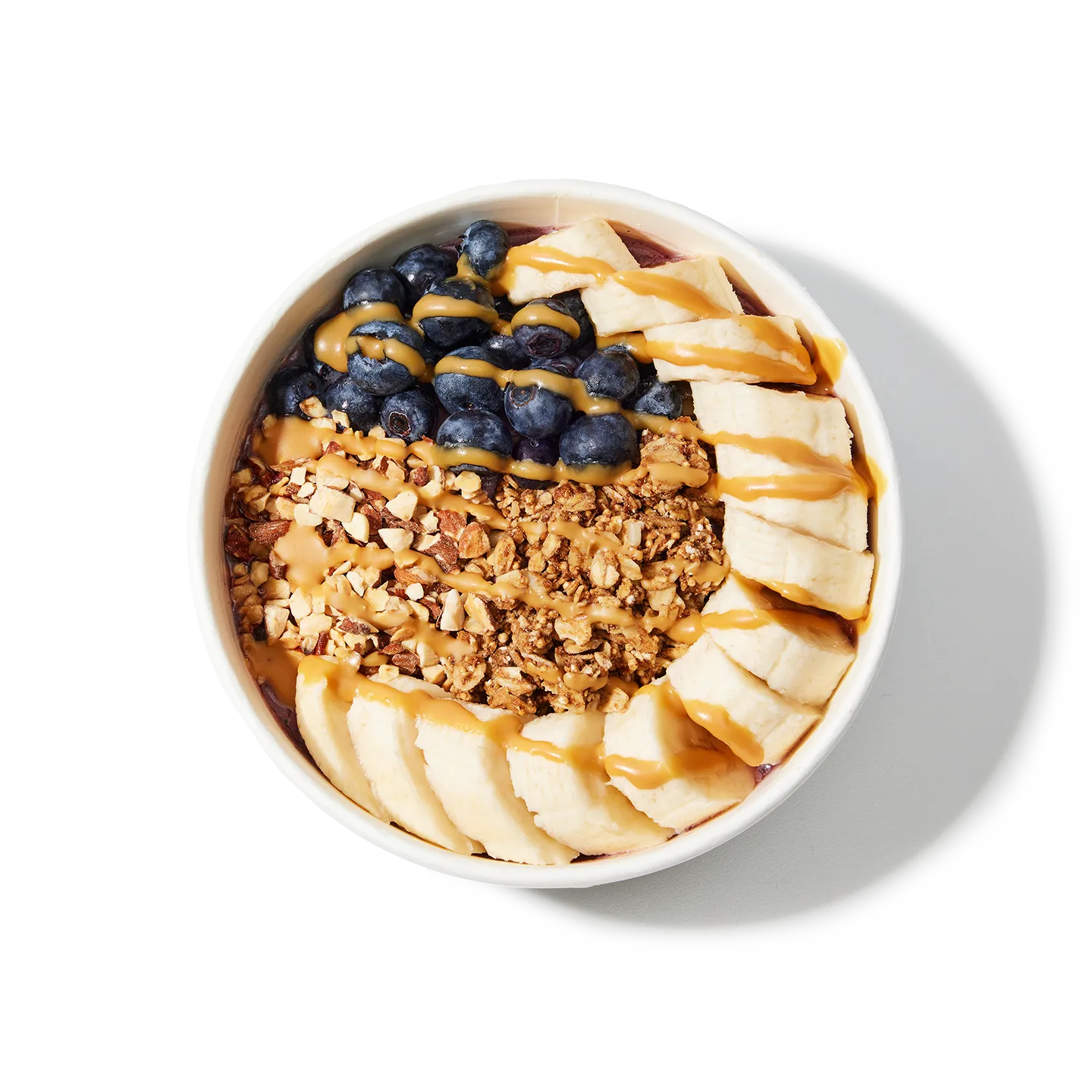 White bowl filled with a layered dessert of granola, sliced bananas, blueberries, and peanut butter on a white background.