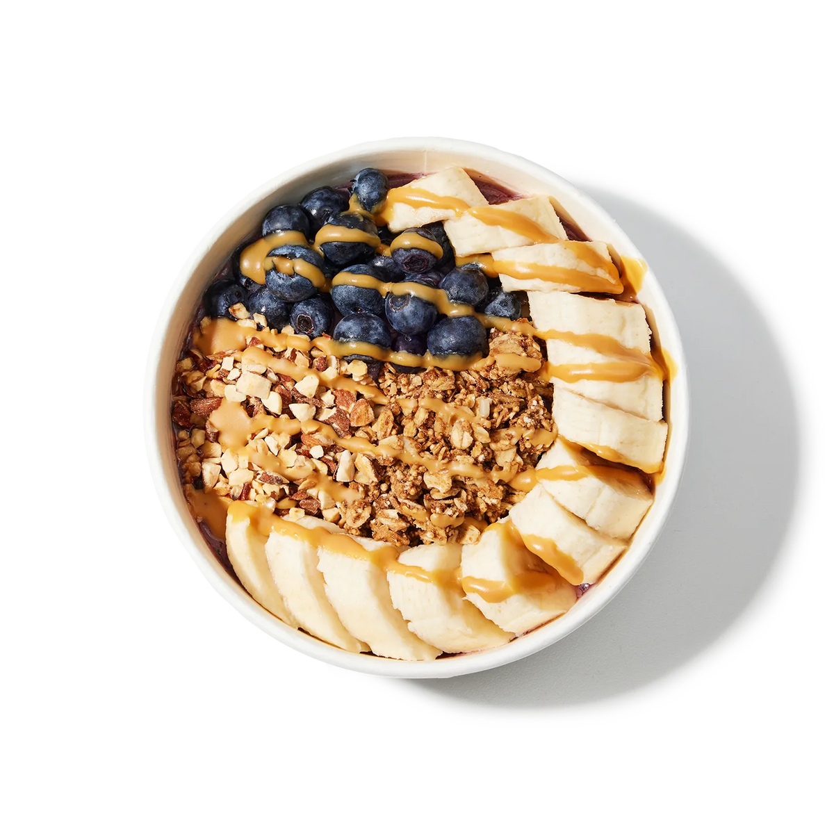 White bowl filled with a layered dessert of granola, sliced bananas, blueberries, and peanut butter on a white background.