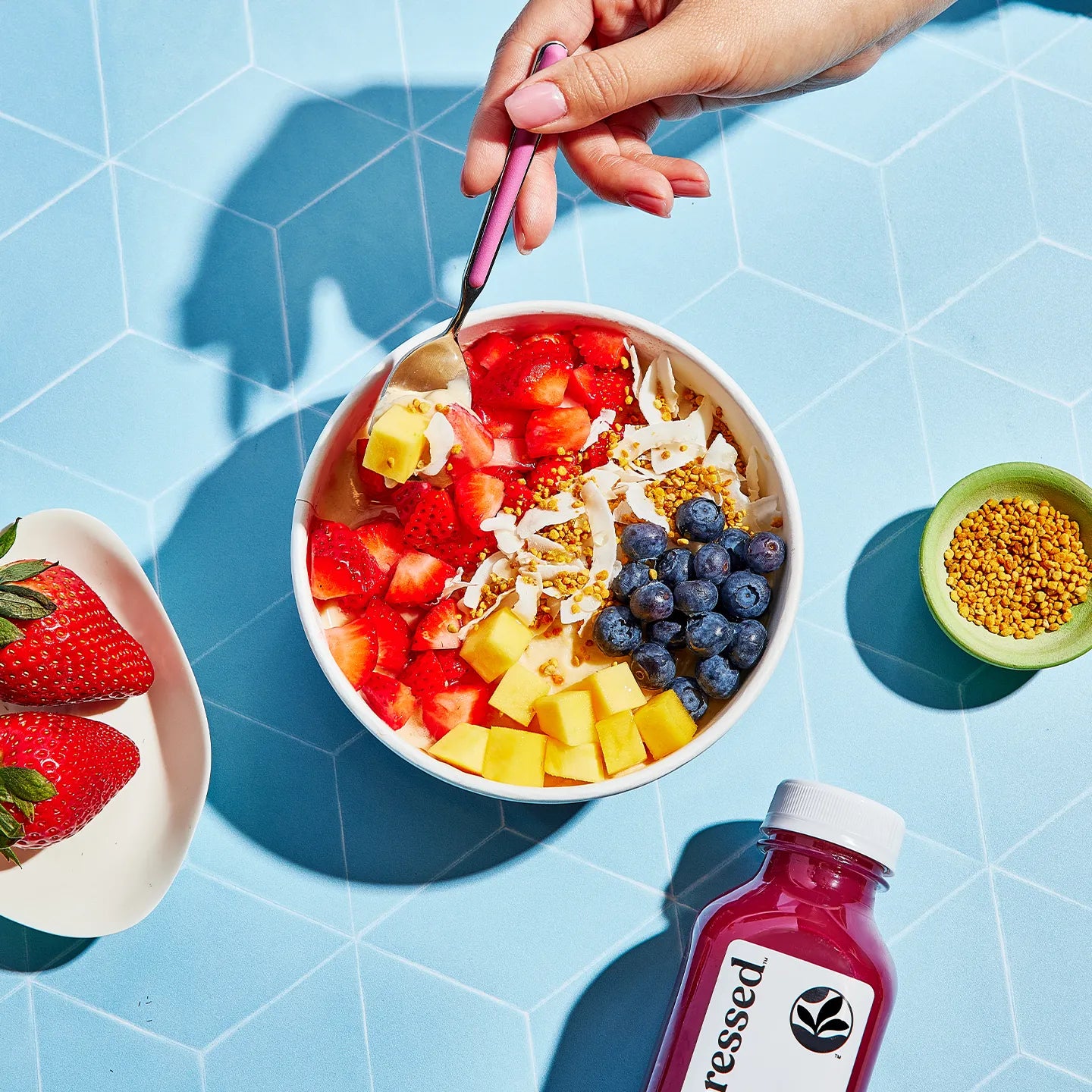 Fruit salad bowl with a spoon, strawberries, blueberries, and a bottle of juice on a blue geometric background.