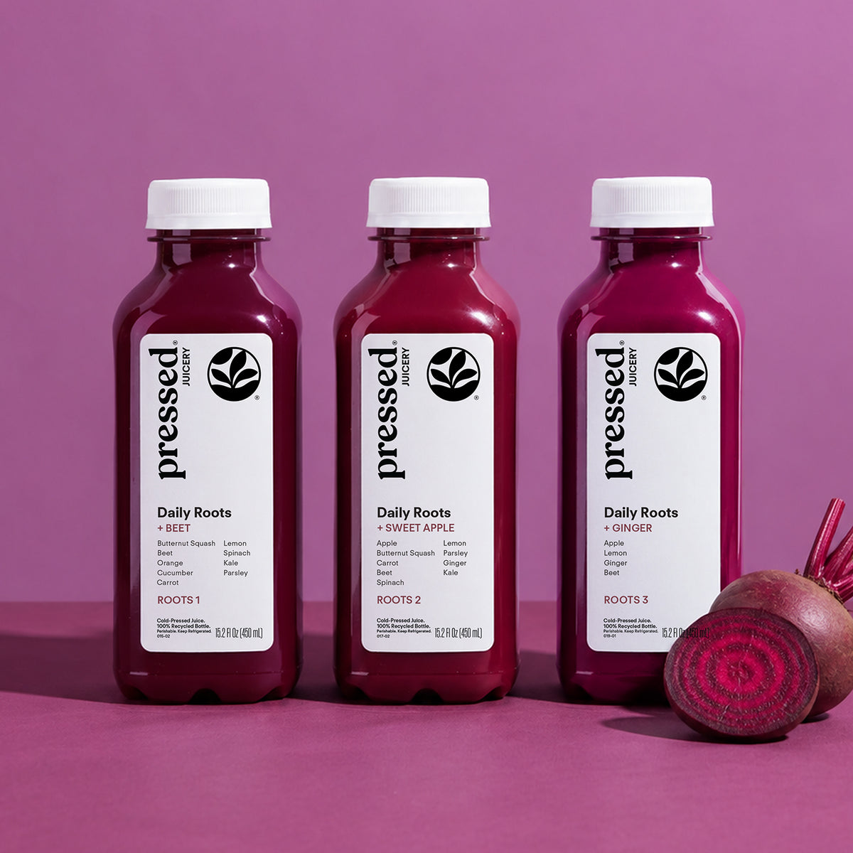 Pressed Juicery Roots 1, Roots 2 + Roots 3 cold-pressed beet juice bottles beside a sliced beet on a pink background.