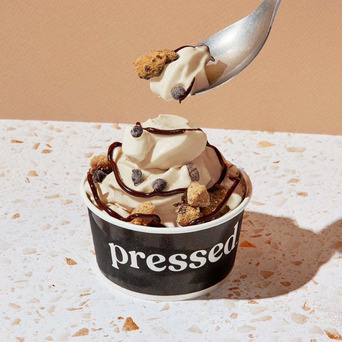 Pressed Juicery Chocolate Chip Sundae with vanilla soft serve, chocolate chips, cookie crumble and cacao drizzle being scooped into with a spoon on a marble countertop
