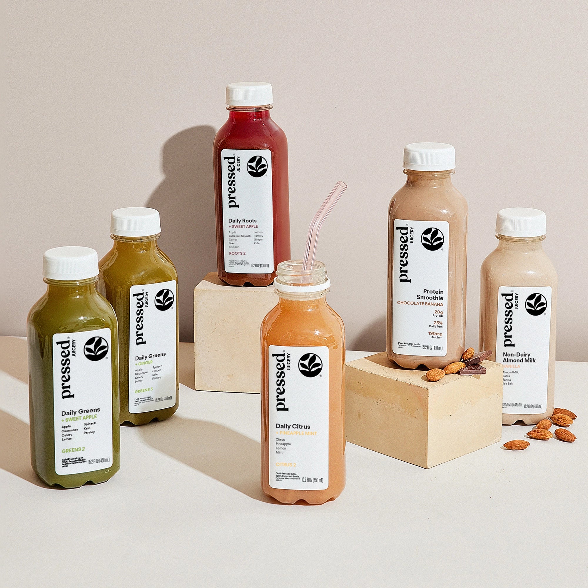 Six bottled juices and drinks from pressed brand arranged on a light beige surface with two wooden blocks. The bottles contain green, yellow-green, red, peach, and beige liquids, each with a white cap and a white label featuring the black pressed logo.