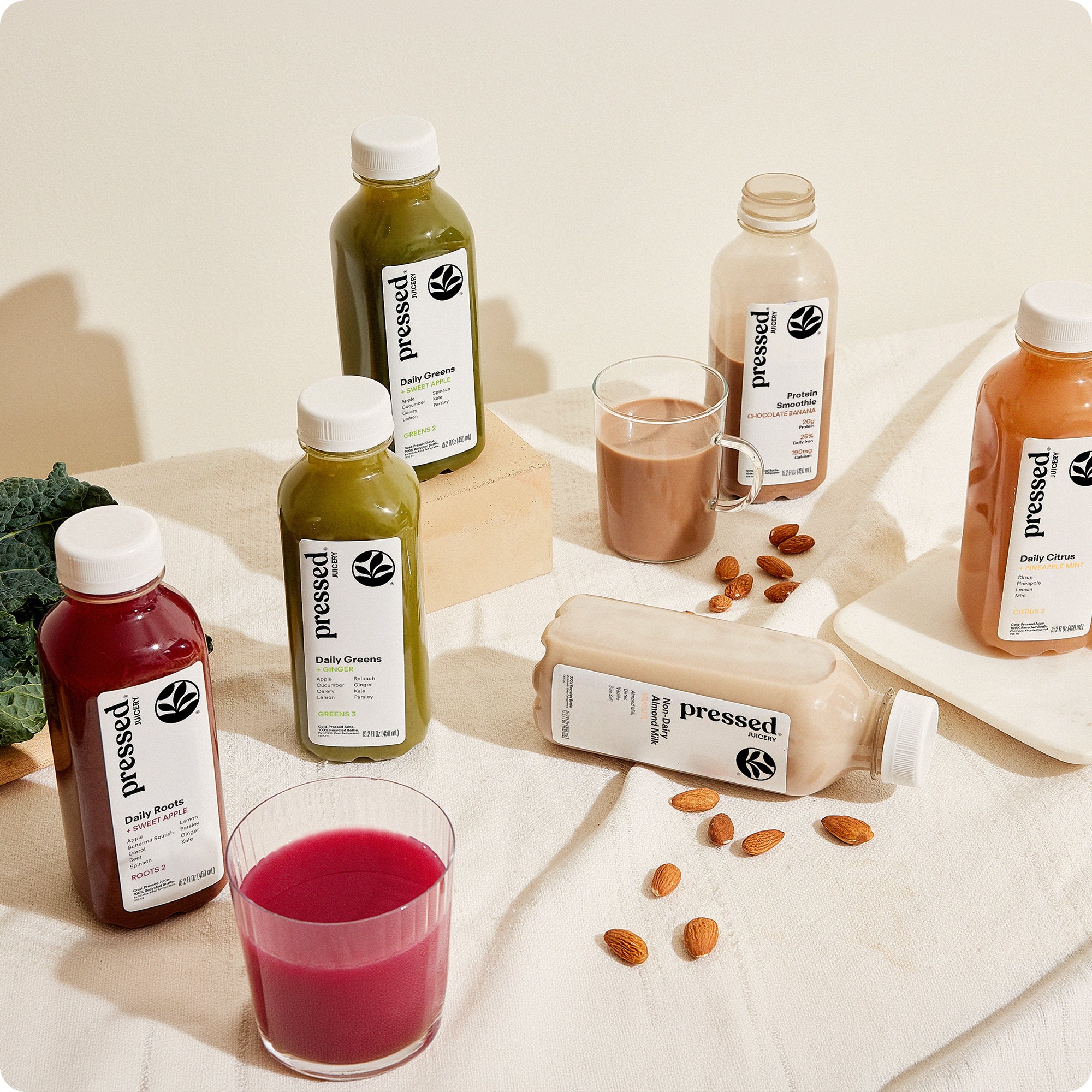 Pressed Juicery Cleanse 1 juice cleanse bottles arranged on a table next to fresh fruit and greenery.