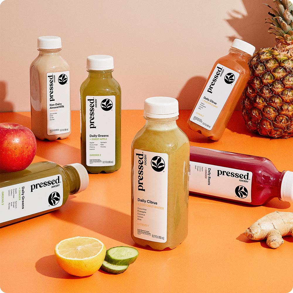 Five bottles of Pressed Juice beverages arranged on an orange surface with a pineapple, kale, lemon, and ginger as props. A black circular badge in the top left reads 'BEST SELLER'.