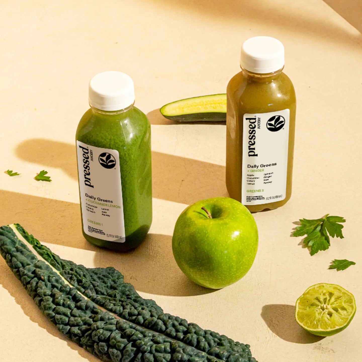Two bottles of 'pressed' green juice with a green apple, kale, and lime on a beige background