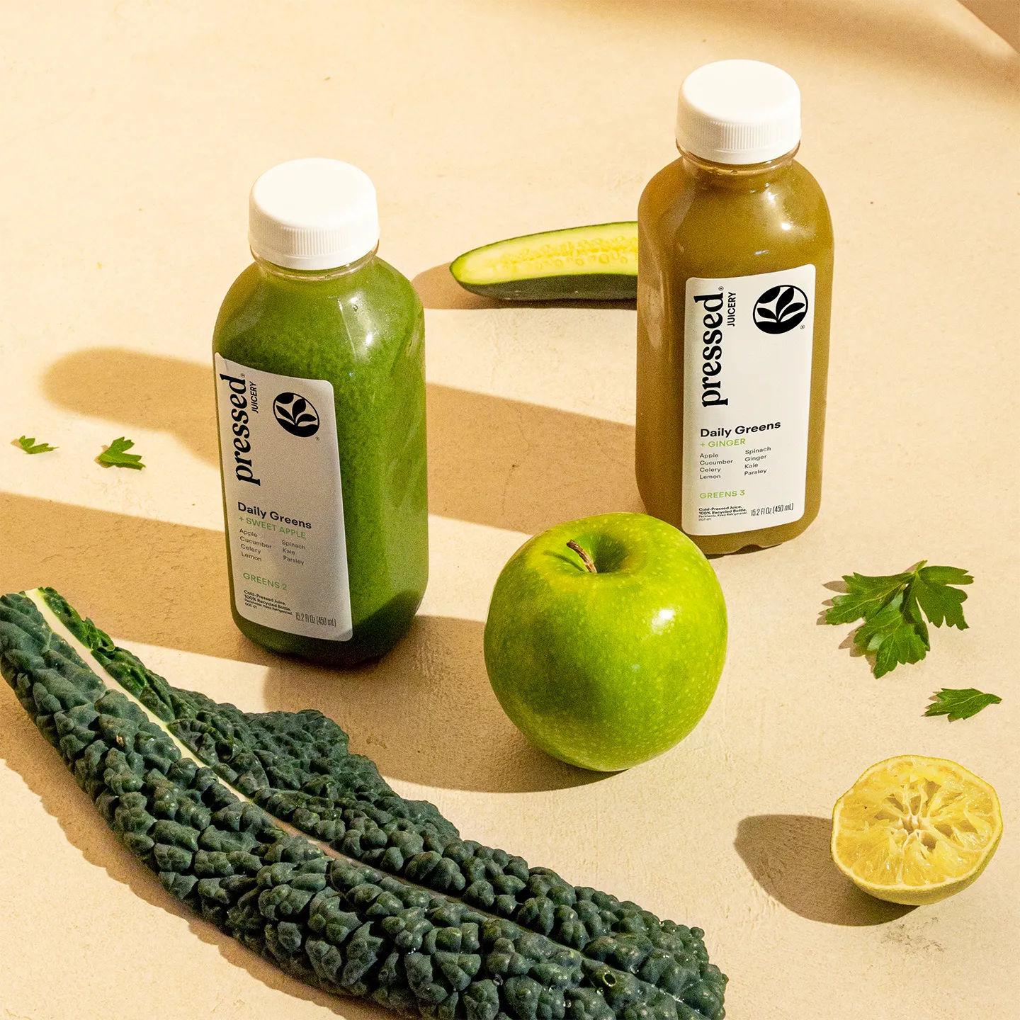 Two bottles of 'pressed' daily greens juice with a green apple, kale, and lemon on a beige background.