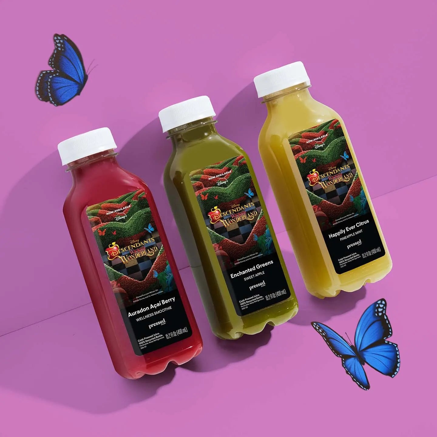 Three plastic juice bottles with white caps are arranged on a pink background with decorative blue butterflies. The bottles contain red, yellow-green, and yellow juices and each has a fantasy label reading 'The Dragon's Breath'.