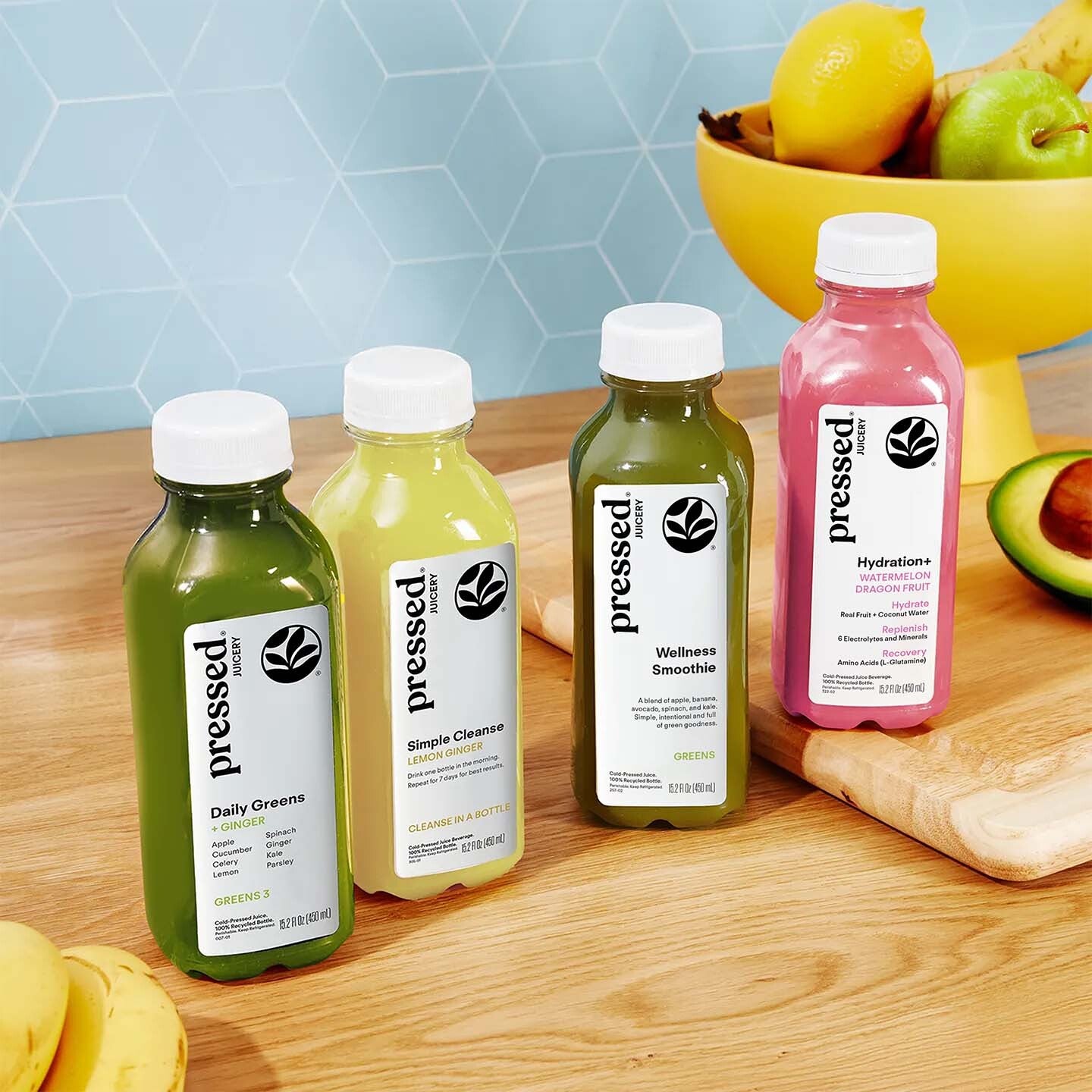 Five 'pressed' juice bottles on a wooden surface with fruits in the background.