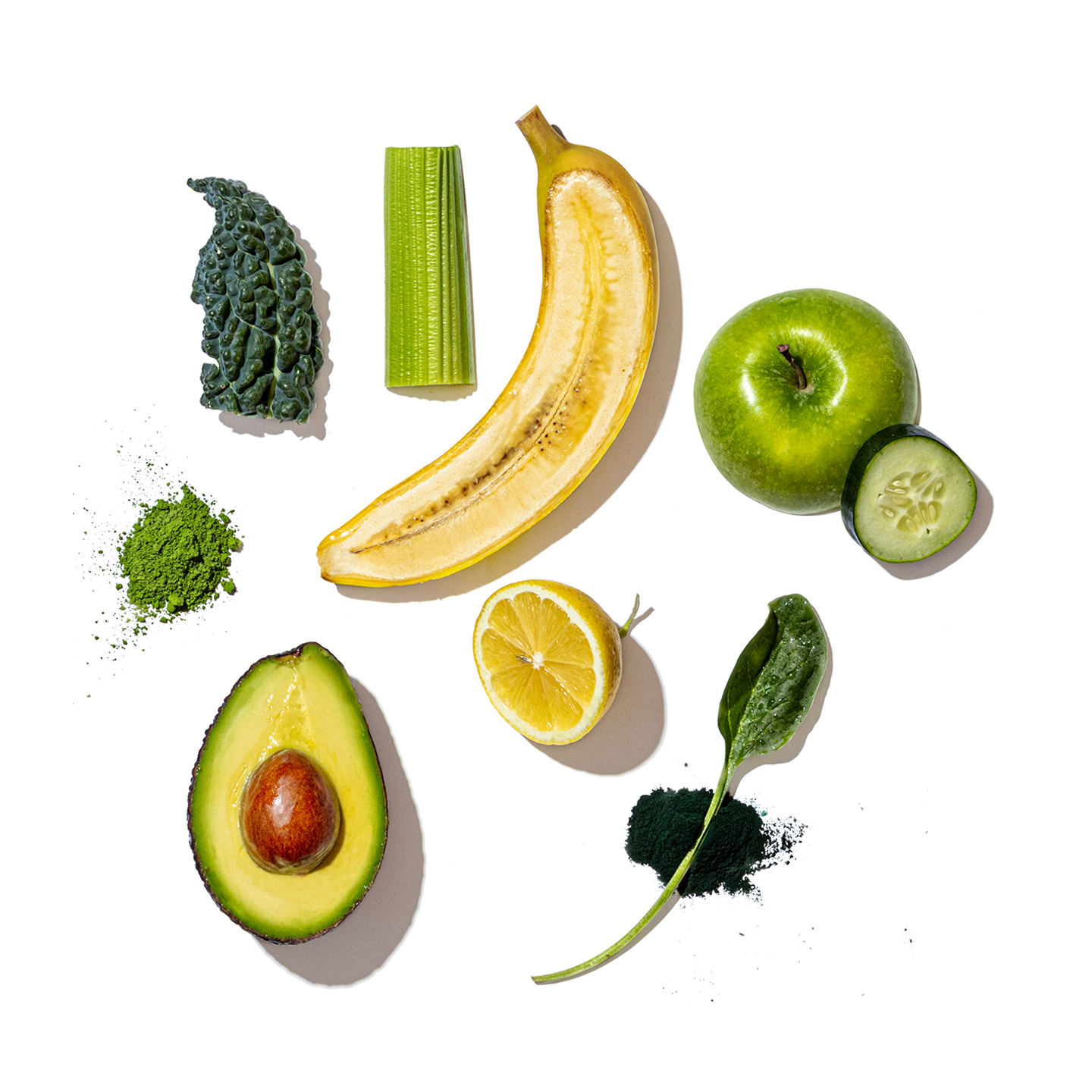 A flat-lay arrangement of green and yellow fruits and vegetables on a white background, including a banana, green apple, cucumber slice, kale, avocado half, lemon half, spinach leaf, and a small pile of dark powder.