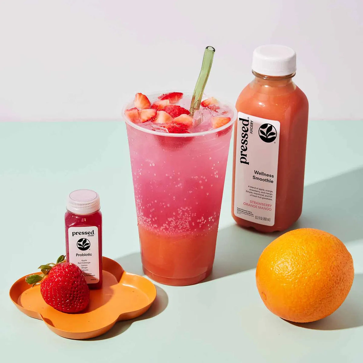 Pink smoothie with strawberries, a bottle of 'pressed' smoothie, a probiotic bottle, an orange, and a strawberry on a light background.