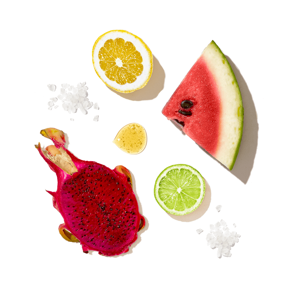 A styled flat-lay of fresh fruits on a white background, including a slice of watermelon, a halved dragon fruit, a halved orange, a lime slice, and a small yellow fruit piece, with scattered coarse salt crystals.