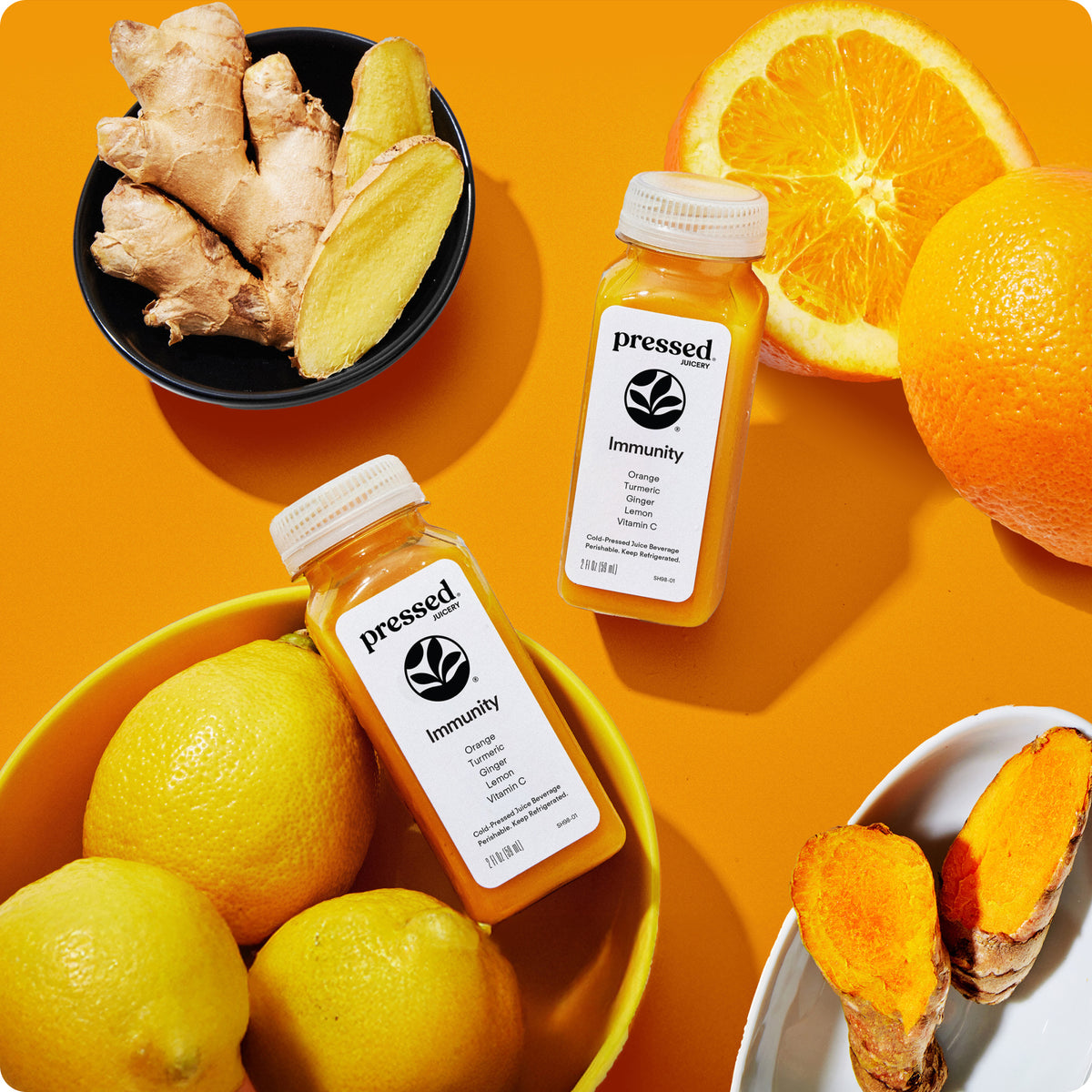 Two Pressed Juicery Immunity Shots with oranges, lemons, ginger and turmeric on an orange background