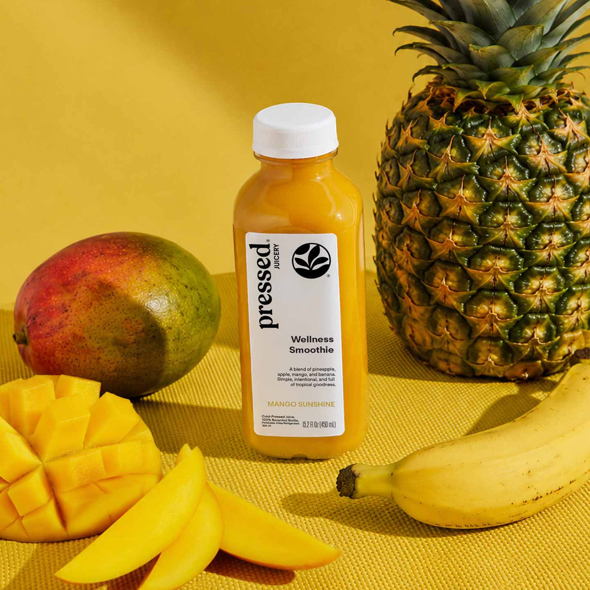 Pressed Juicery Mango Sunshine Wellness Smoothie bottle surrounded by fresh mango, pineapple and banana on a yellow background.
