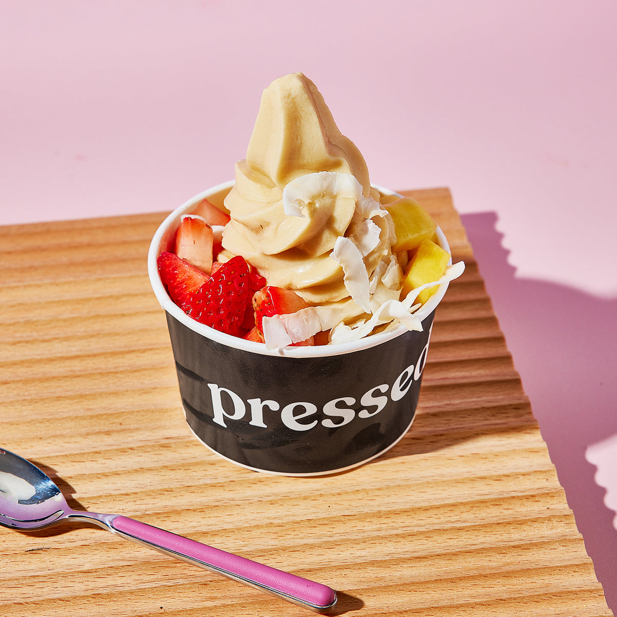 Pressed Juicery Tropical Sundae with tropical soft serve, strawberries, mango and coconut flakes on a table next to a pink wall and pink spoon