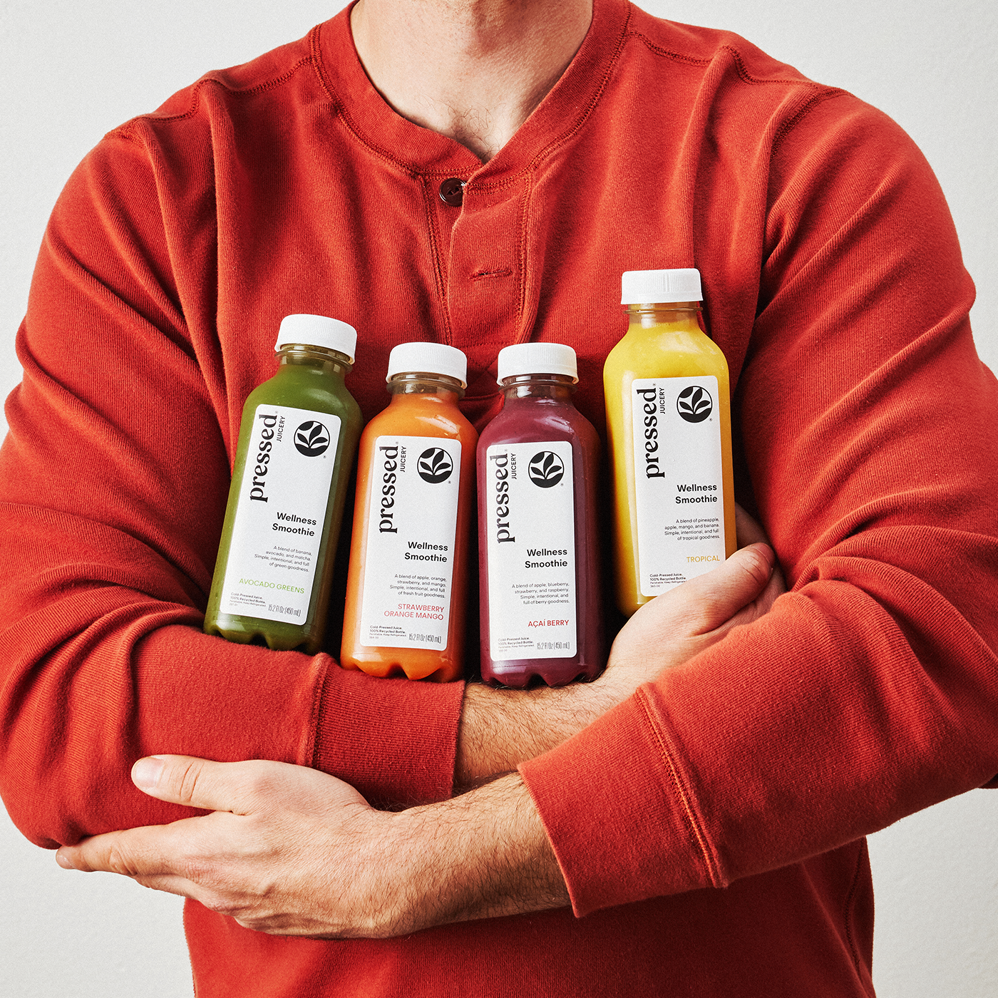 A person in a rust-red long-sleeve henley shirt holds four bottled juices against their chest. The bottles are labeled 'pressed' and contain green, orange, pinkish-red, and yellow liquids.