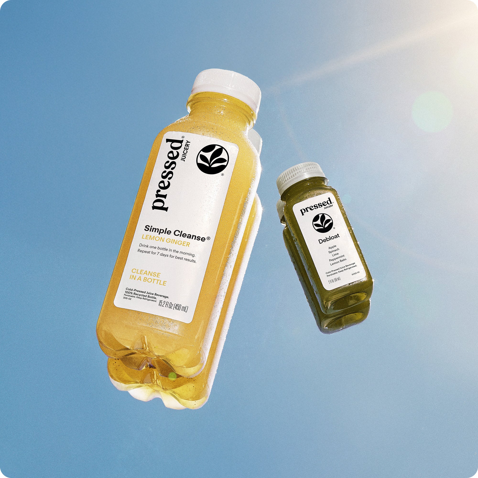 Two bottles of Pressed Juices, Simple Cleanse Juice and Debloat Shot, on a mirror reflecting a blue sky.