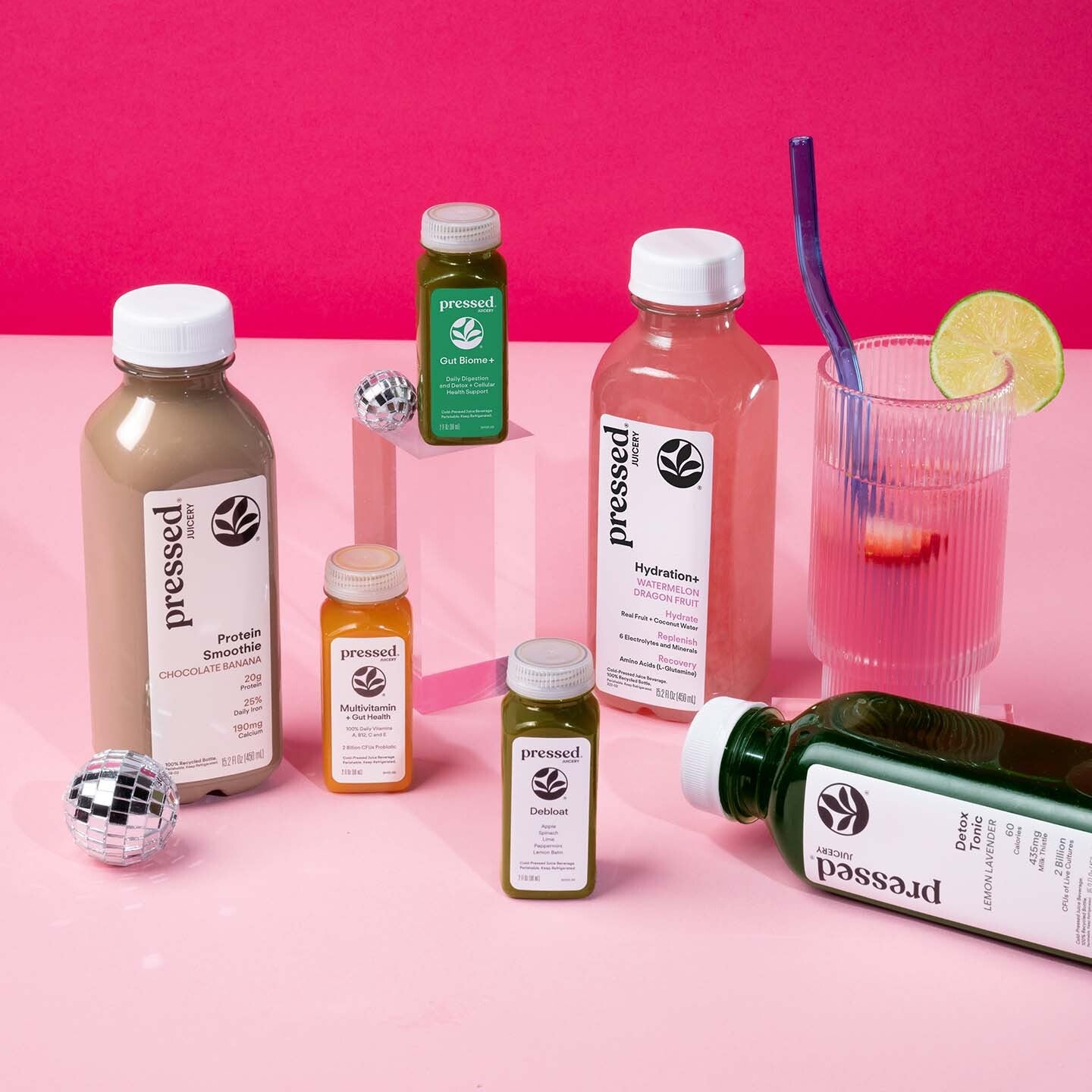 A styled product shot of multiple Pressed brand bottled drinks and a glass on a pink surface against a hot pink background. Visible products include a large protein smoothie bottle, a hydration drink bottle, smaller juice bottles, and a glass with a purple straw and lime wedge, plus disco ball props.