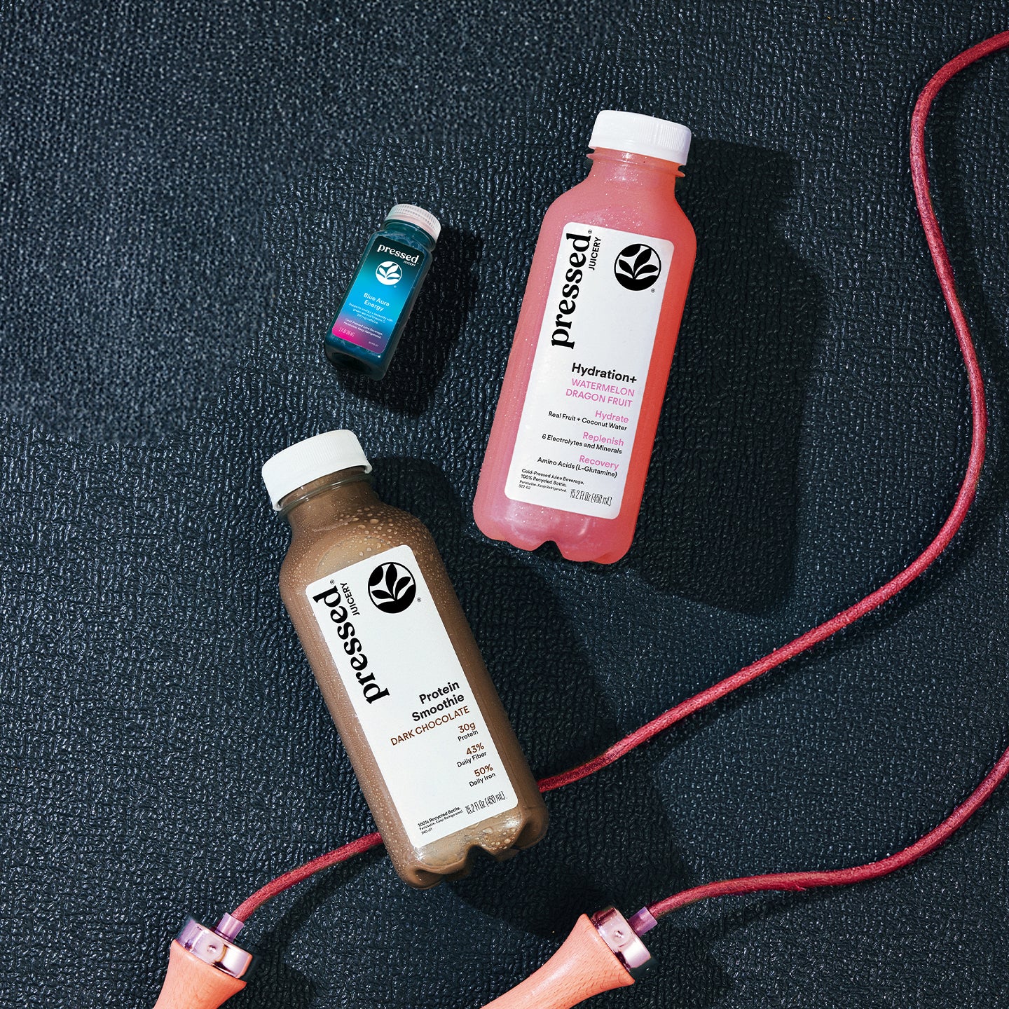 Pressed Juicery Workout Essentials Pack featuring a Dark Chocolate Protein Smoothie, Hydration+ Watermelon Dragon Fruit drink, and Blue Aura Energy shot with a jump rope.