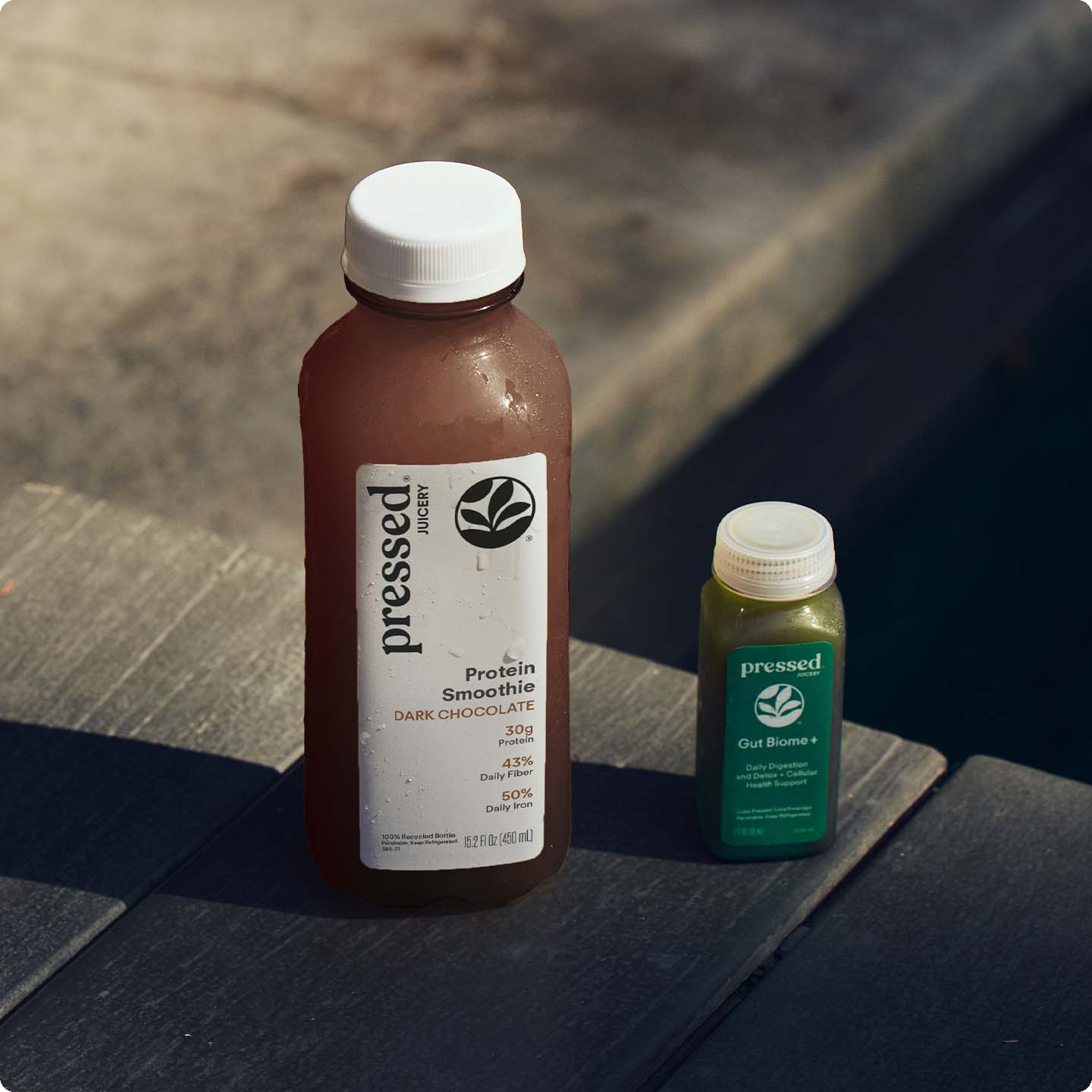 Two bottles of Pressed Foods protein powder on a dark wooden table in natural sunlight. The larger bottle on the left is a brown Protein Smoothie Dark Chocolate flavor, and the smaller bottle on the right is a green Our Biotics+ powder.