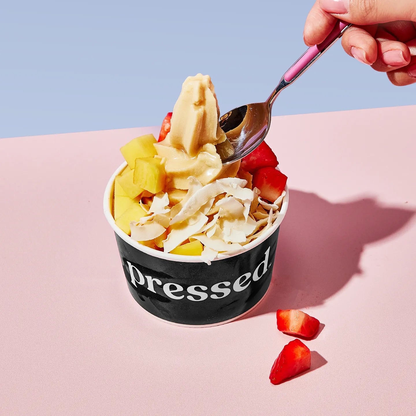 Pressed Juicery Tropical Protein Freeze with fruit on a pink surface against a blue background.