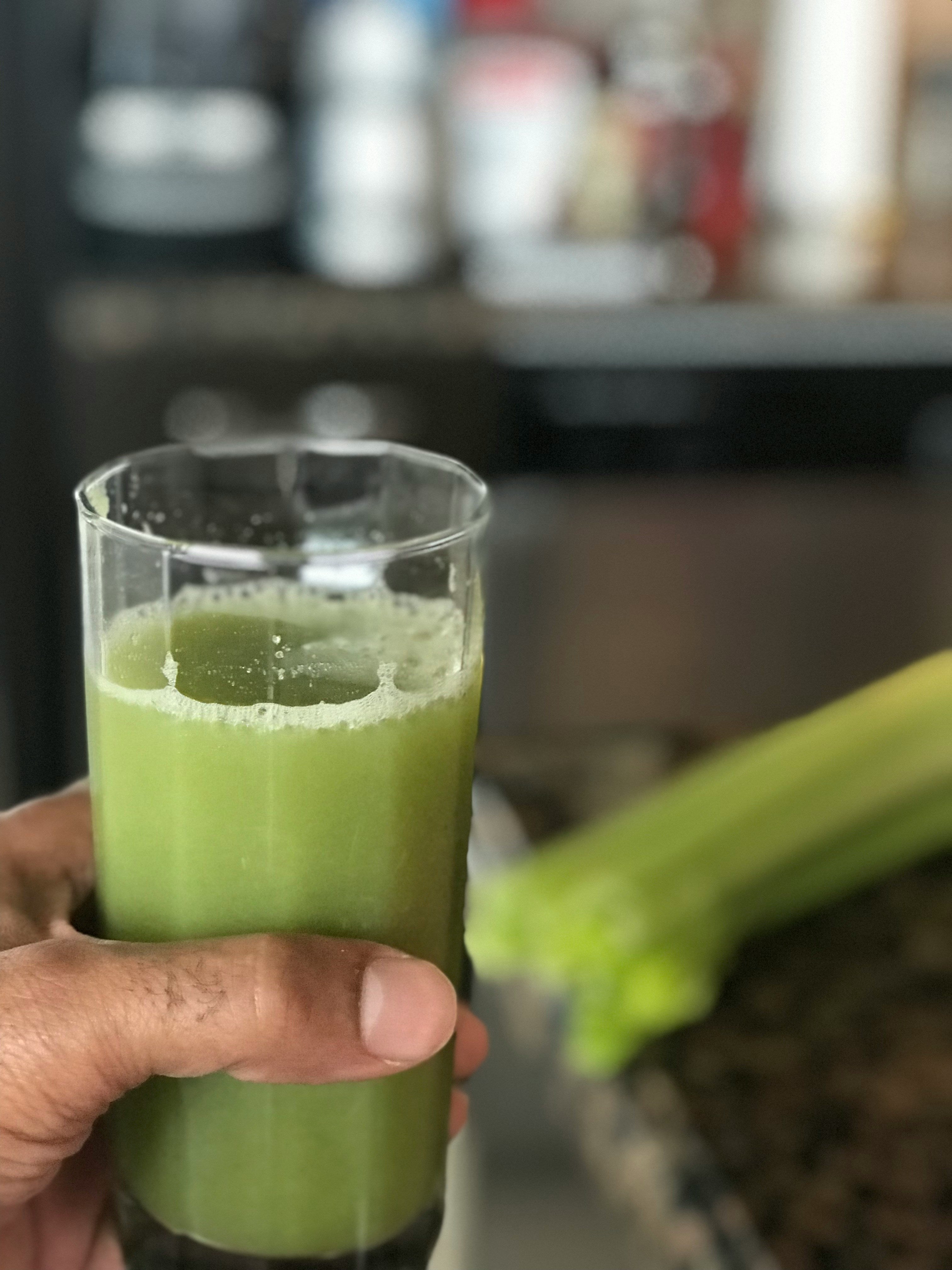An image of a hand holding a glass of green juice with celery in the background