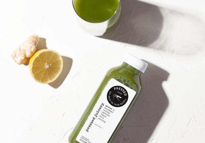 Bottle of pressed green juice next to a lemon and some ginger on a white background.