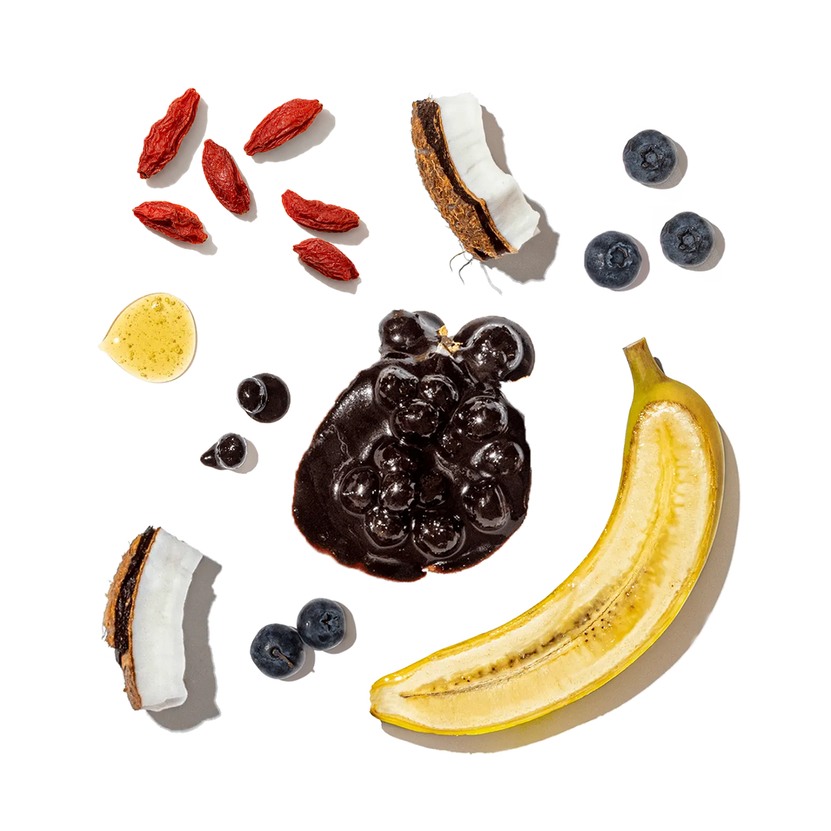Fruits and ingredients including a banana, blueberries, goji berries, a coconut, and honey on a white background.