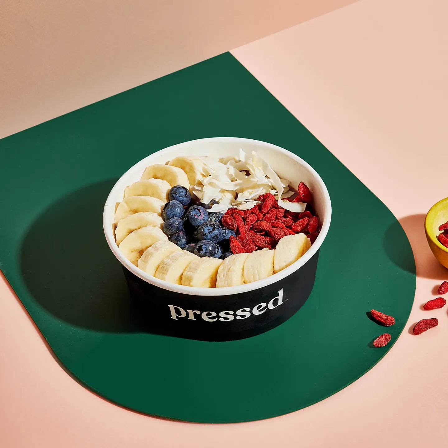 Fruit bowl with bananas, blueberries, and goji berries in a container labeled 'pressed' on a green and beige background.