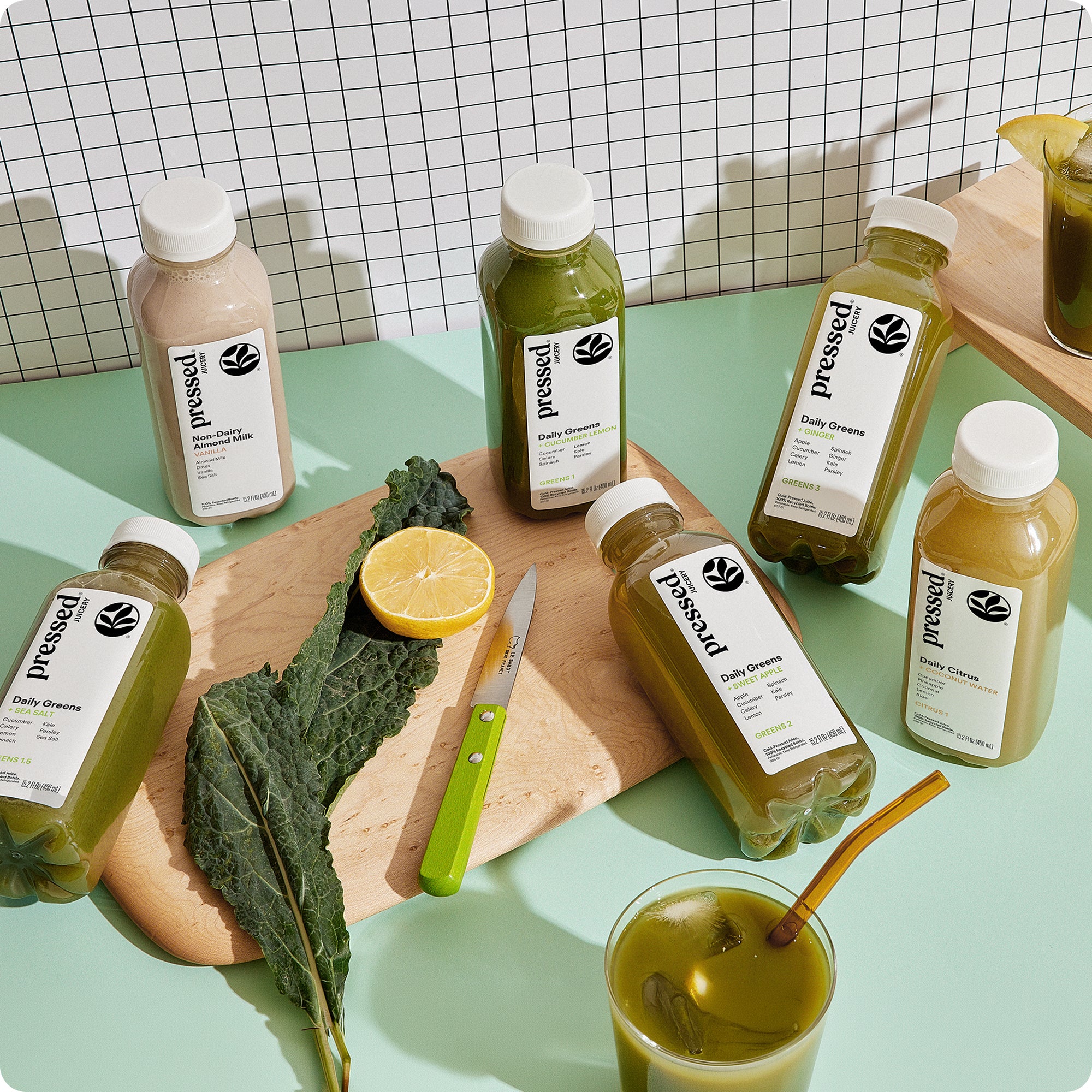 Pressed Juicery Cleanse 3 juice cleanse bottles arranged on a table next to kale, lemon and fresh greenery.