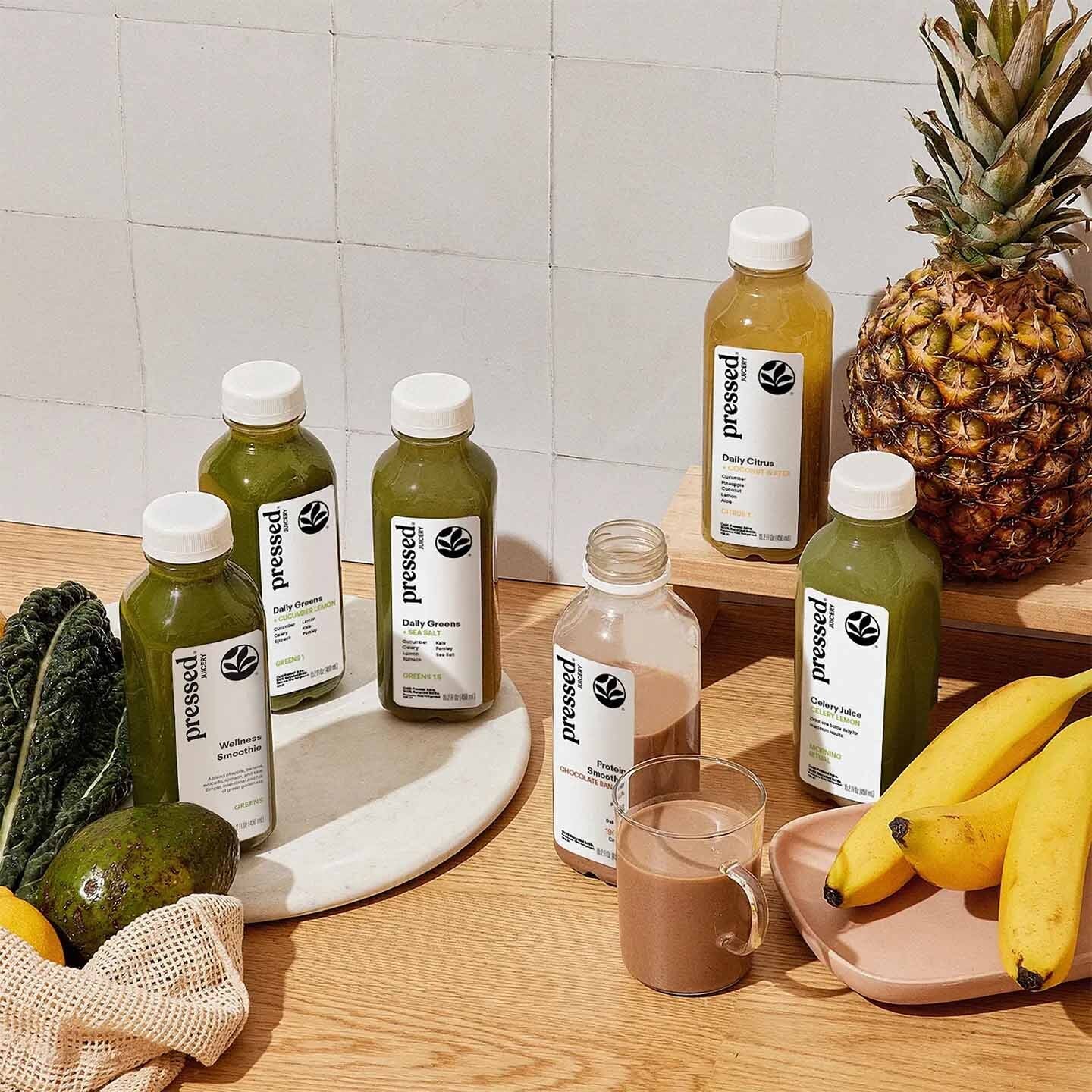 Six bottles of Pressed Juice beverage arranged on a wooden table with a white tiled wall behind them, alongside fruits including a pineapple, bananas, kale, avocado, and a glass of mixed drink. No people or animals are present.