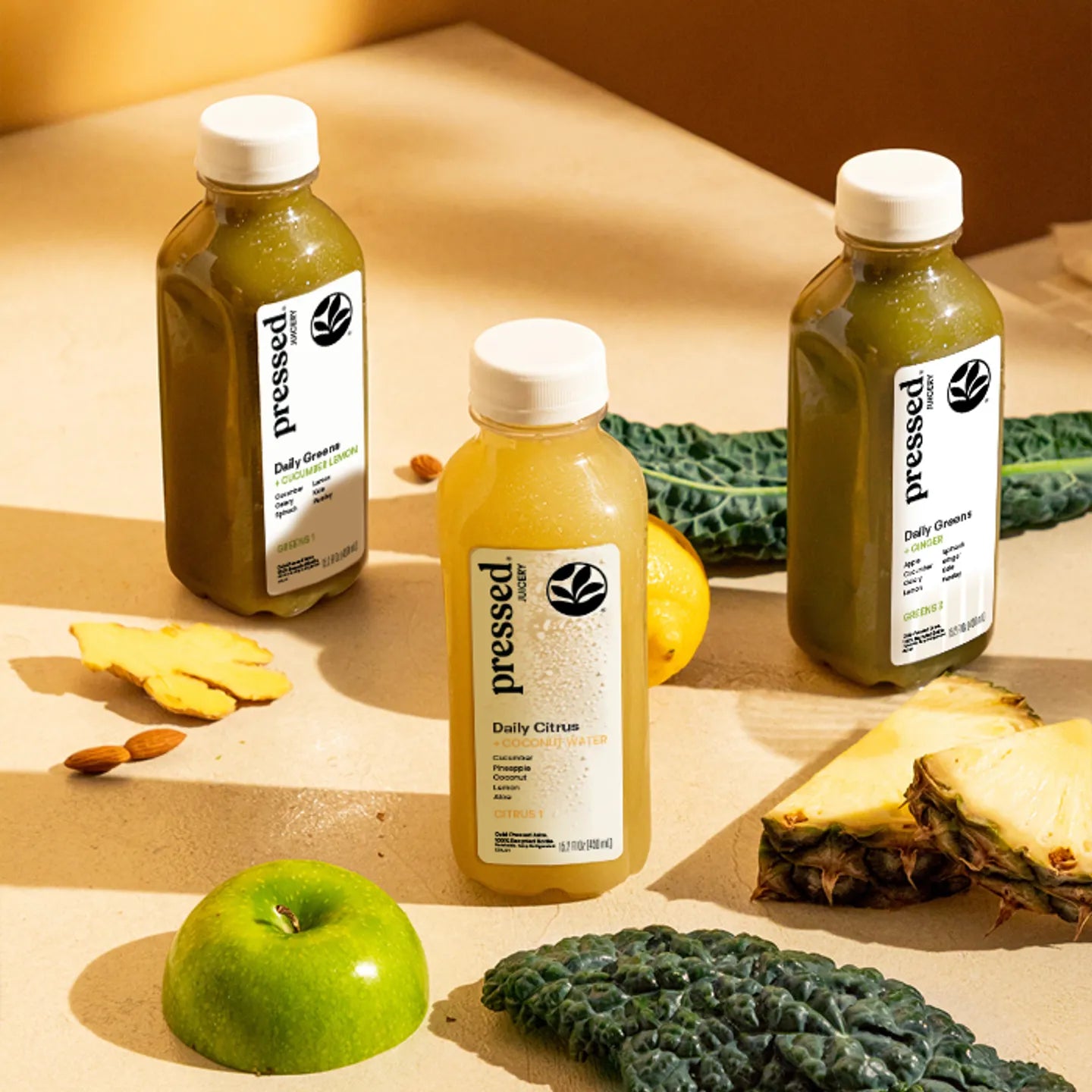 Three bottles of 'pressed' daily juice on a wooden surface with fruits and vegetables.