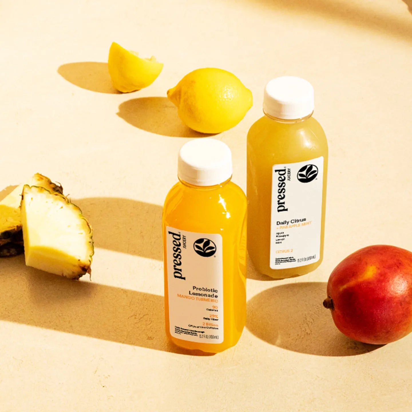 Two bottles of 'Pressed' juice with lemons, a pineapple, and an apple on a beige background
