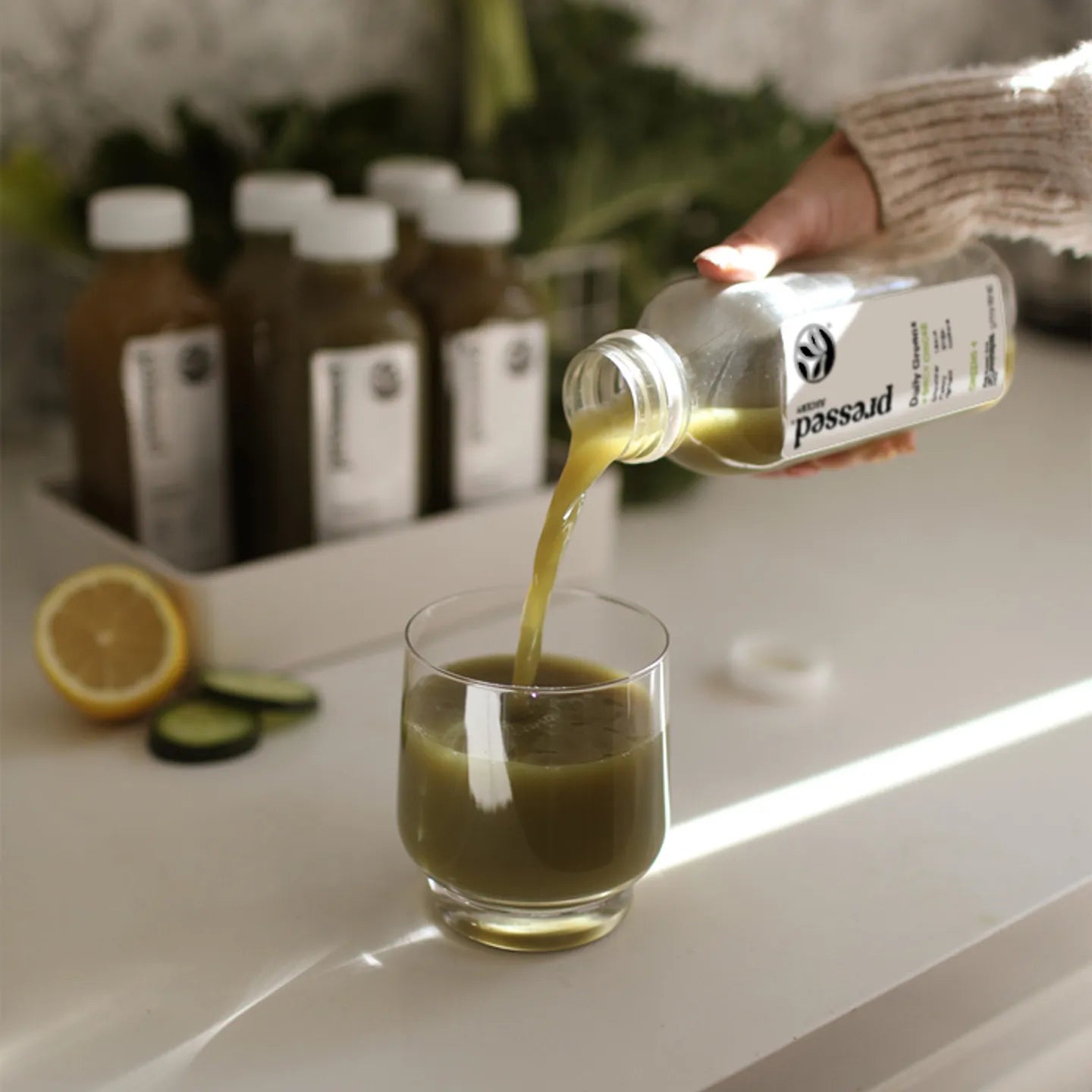 Person pouring green juice from a bottle into a glass with bottles and fruits in the background.