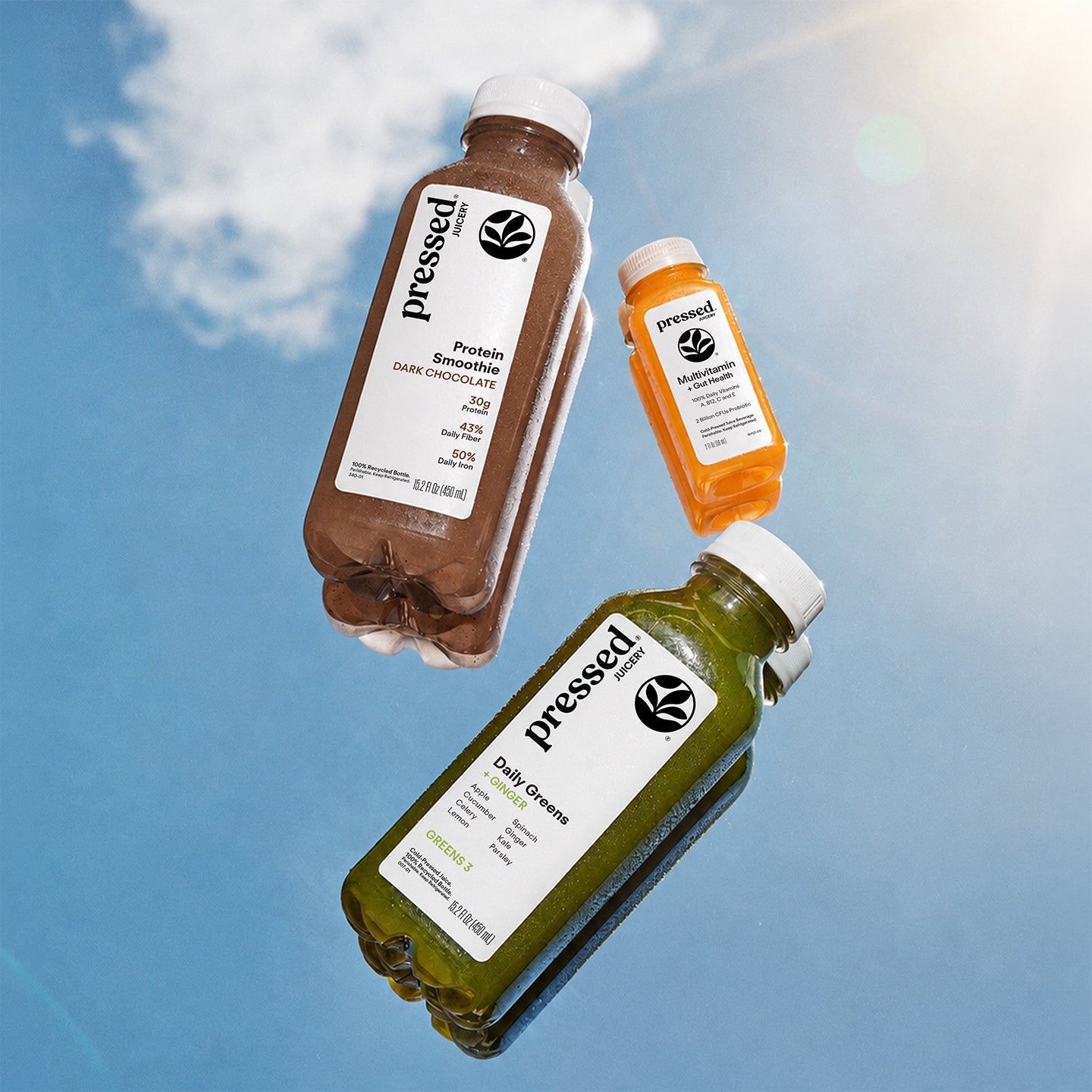 Three plastic bottles of Pressed brand liquid supplements floating against a blue sky background with clouds and sun rays. The bottles contain dark brown, orange, and green liquids and have white caps and white labels with black text.