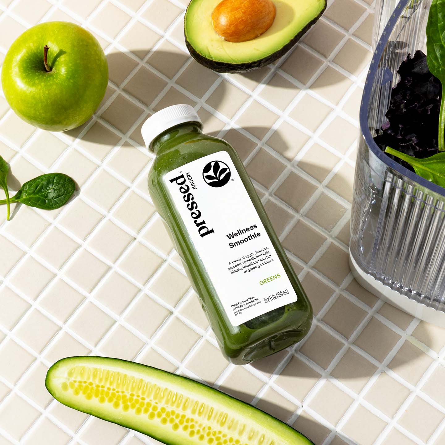 A bottle of pressed wellness smoothie green juice lies on a white tiled surface, surrounded by fresh ingredients including a green apple, avocado, cucumber, and a glass blender jar.