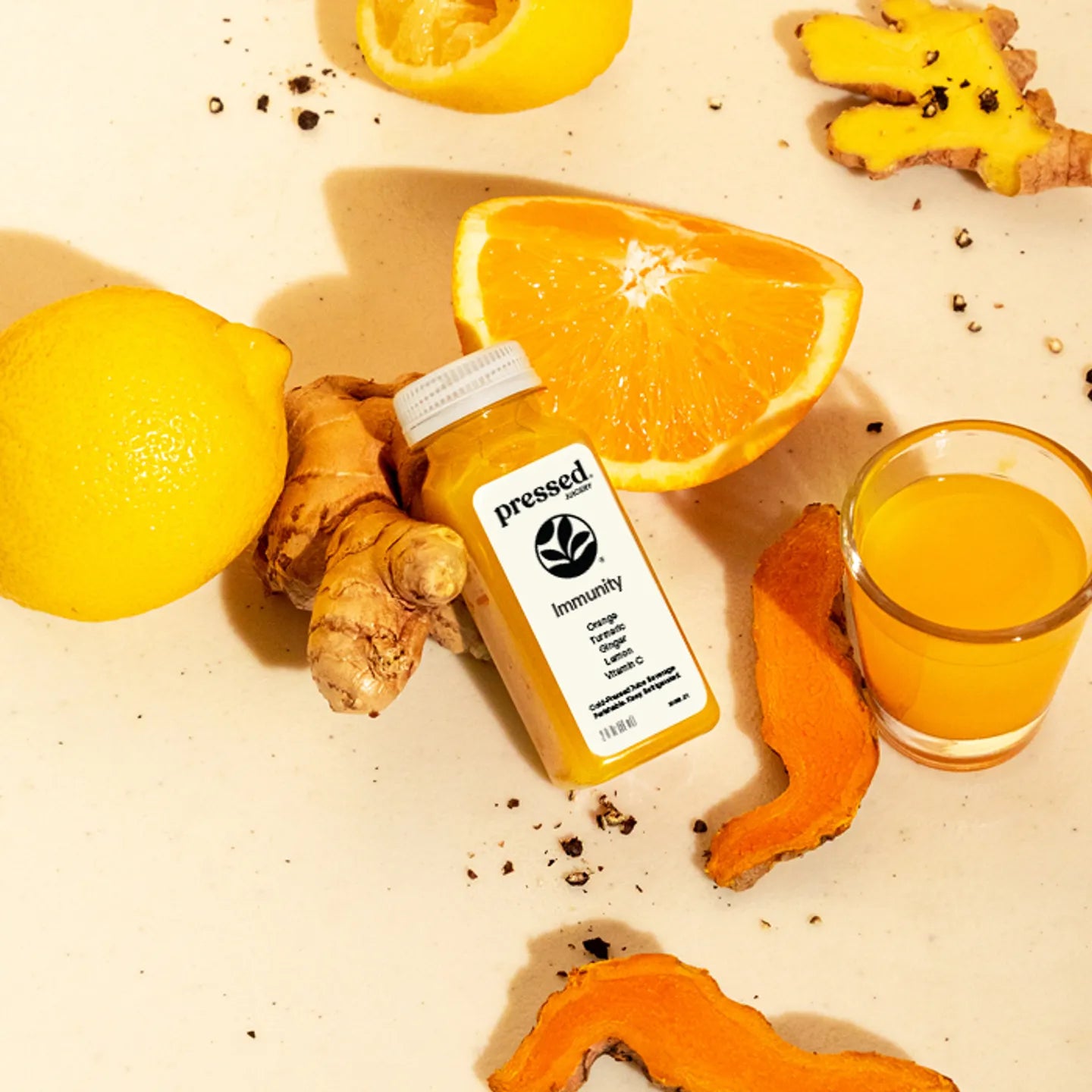 Bottle of 'Pressed' immunity supplement with lemons, oranges, ginger, and a glass of juice on a beige surface.