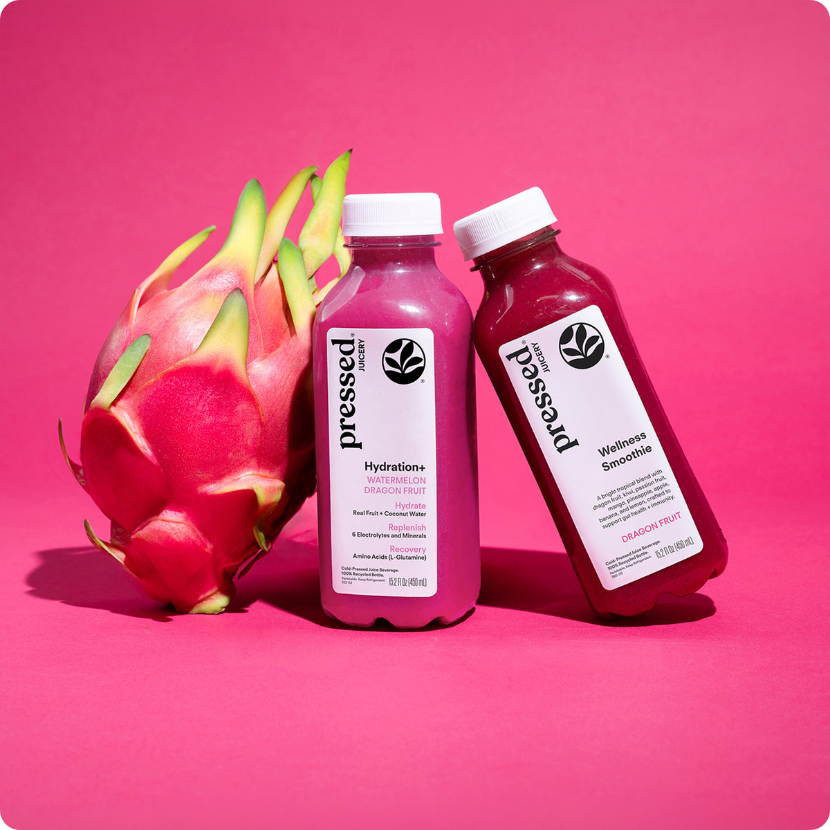 Pressed Hydration+ Watermelon Dragon Fruit and Dragon Fruit Wellness Smoothie bottles beside fresh dragon fruit on a bright pink background.