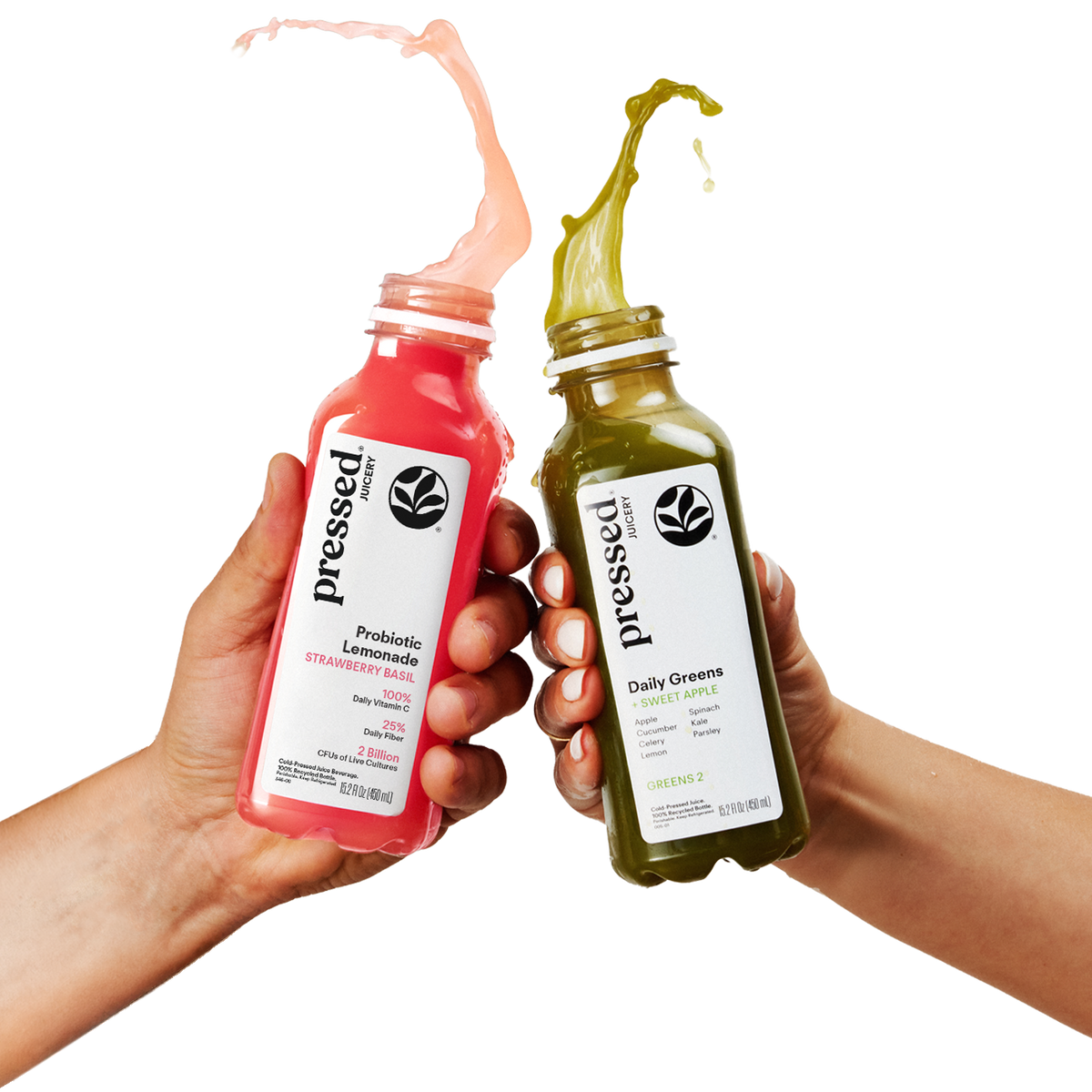 Two hands holding pressed brand bottled juices, one pink strawberry lemonade and one green daily greens, with liquid splashing from the open tops against a white background.