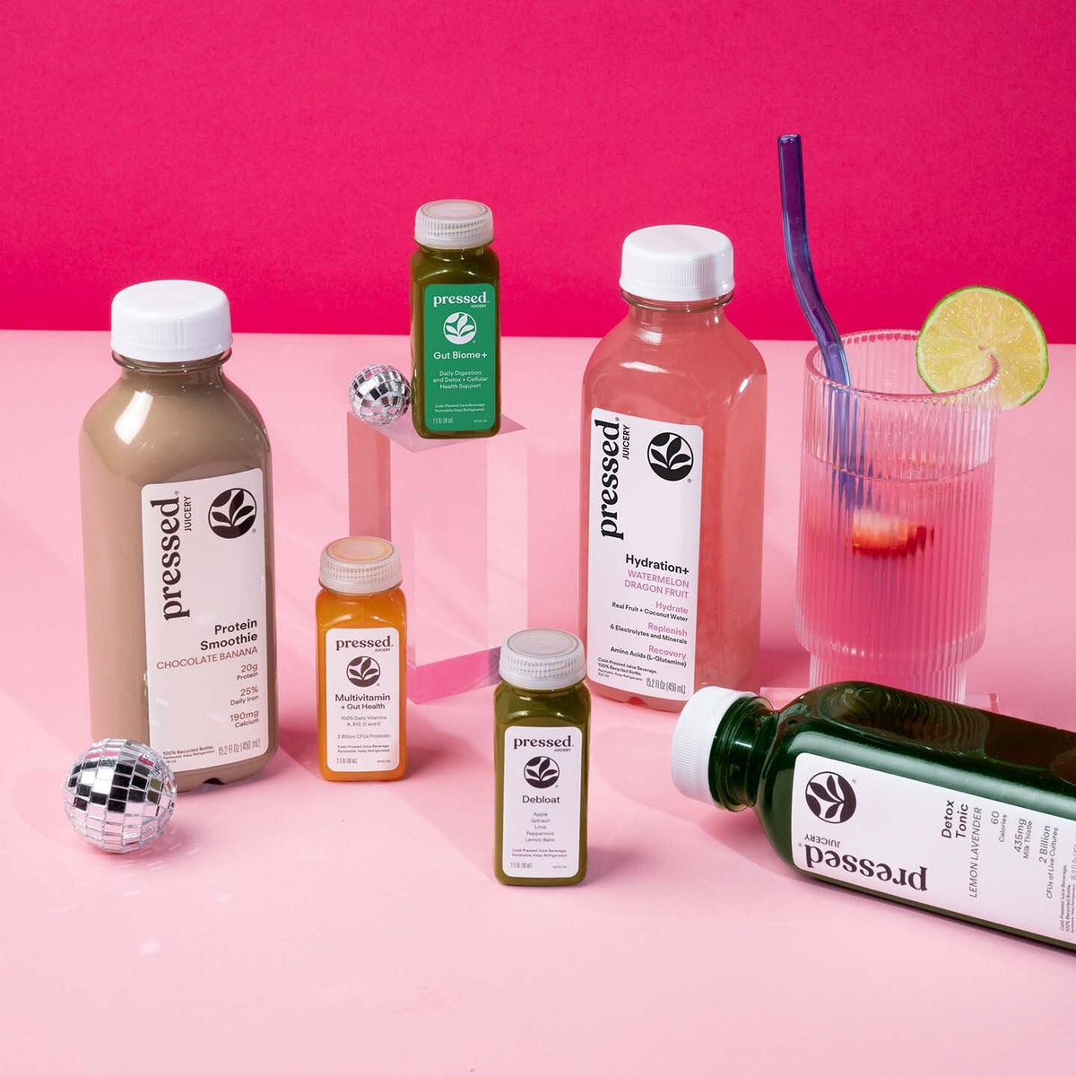 A styled product shot of multiple Pressed brand bottled drinks and a glass on a pink surface against a hot pink background. Visible products include a large protein smoothie bottle, a hydration drink bottle, smaller juice bottles, and a glass with a purple straw and lime wedge, plus disco ball props.