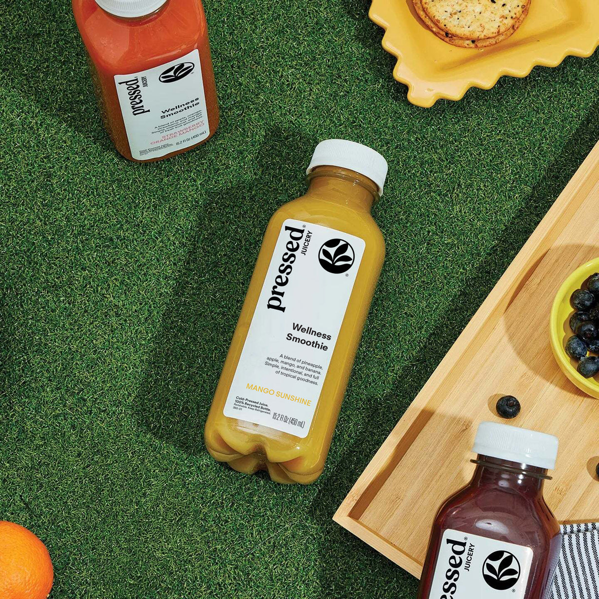Bottles of pressed wellness smoothie on a green surface with fruits and a wooden board.