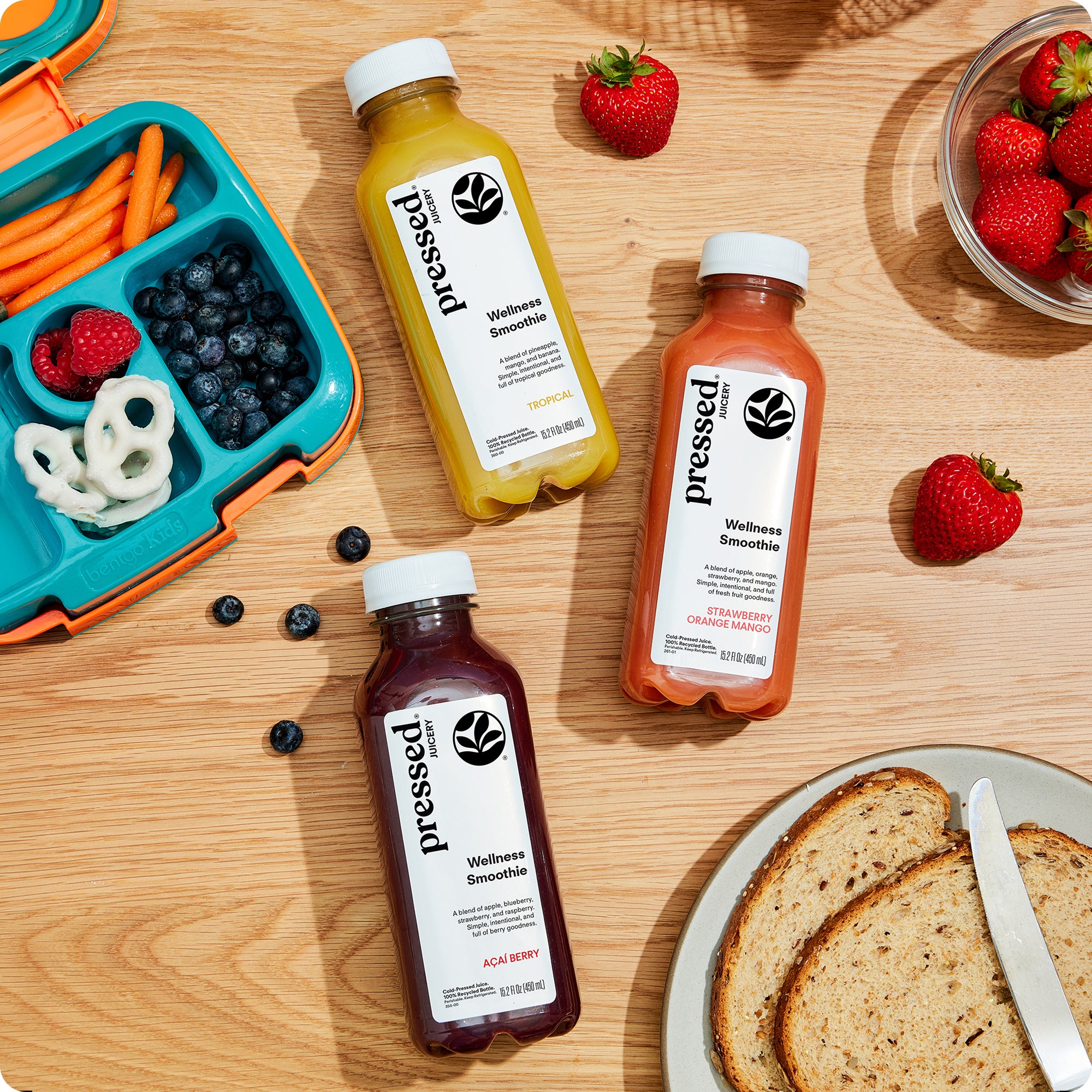 Three bottles in the Pressed Wellness Smoothie Pack arranged on a wooden table with a lunchbox, strawberries, blueberries, and toast nearby.