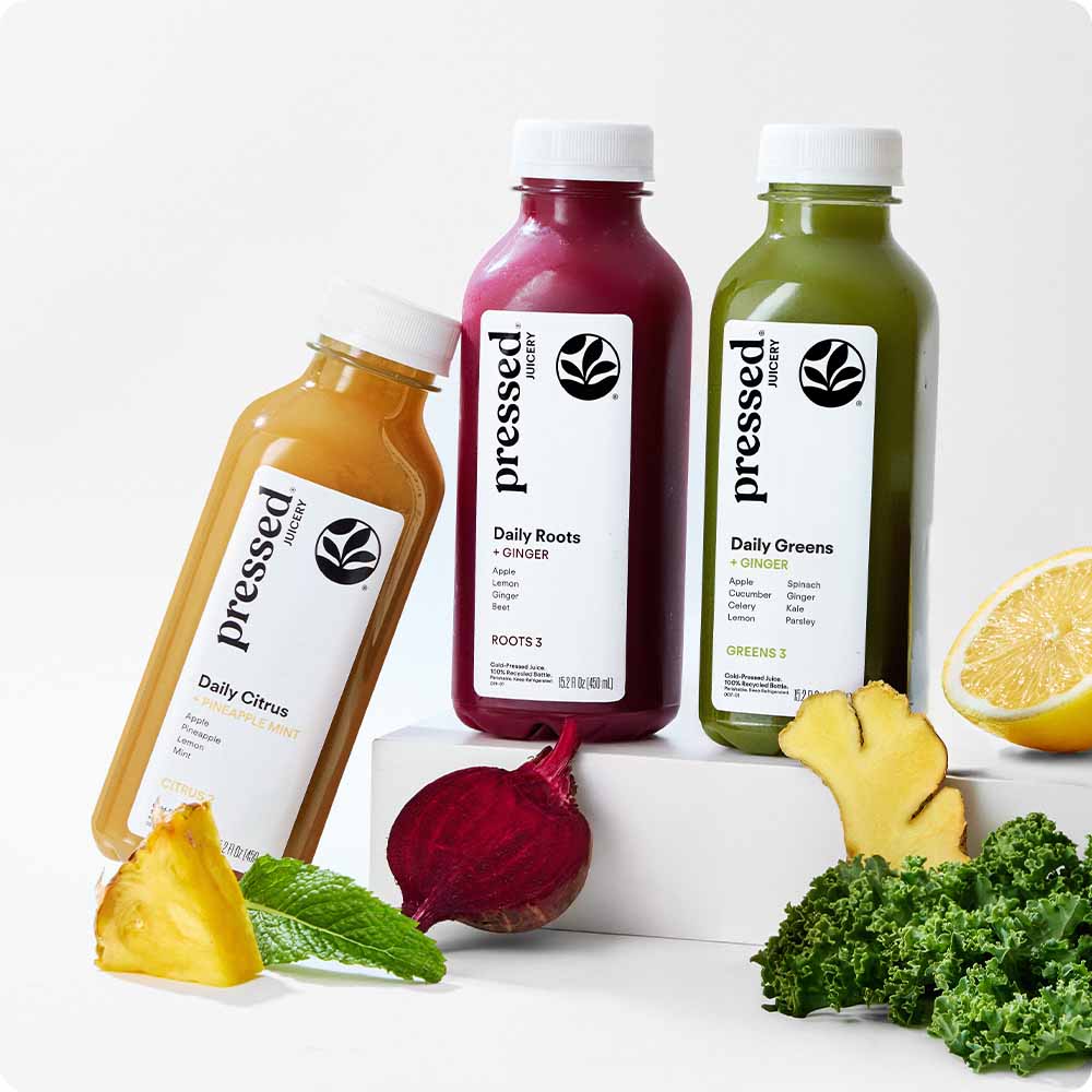 Three bottled juices from pressed fruit are arranged on white blocks with fresh produce around them: an orange Daily Citrus bottle leaning left, a magenta Daily Roots + Ginger bottle standing center, and a green Daily Greens bottle standing right. Pineapple, beet, kale, and lemon slices are visible in the scene.