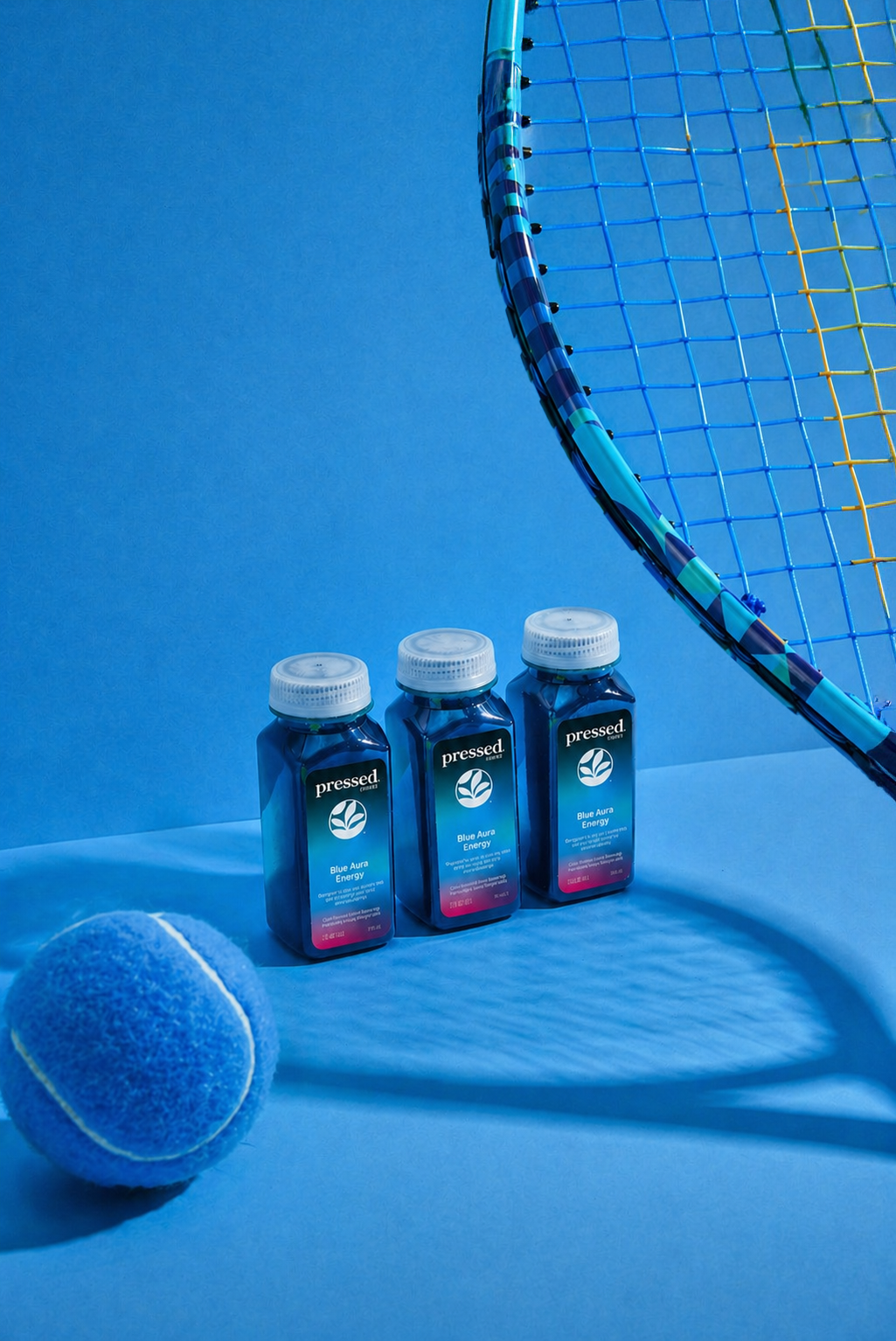 Three bottles of Blue Aura Energy Shots on a blue background with a tennis ball and racket 