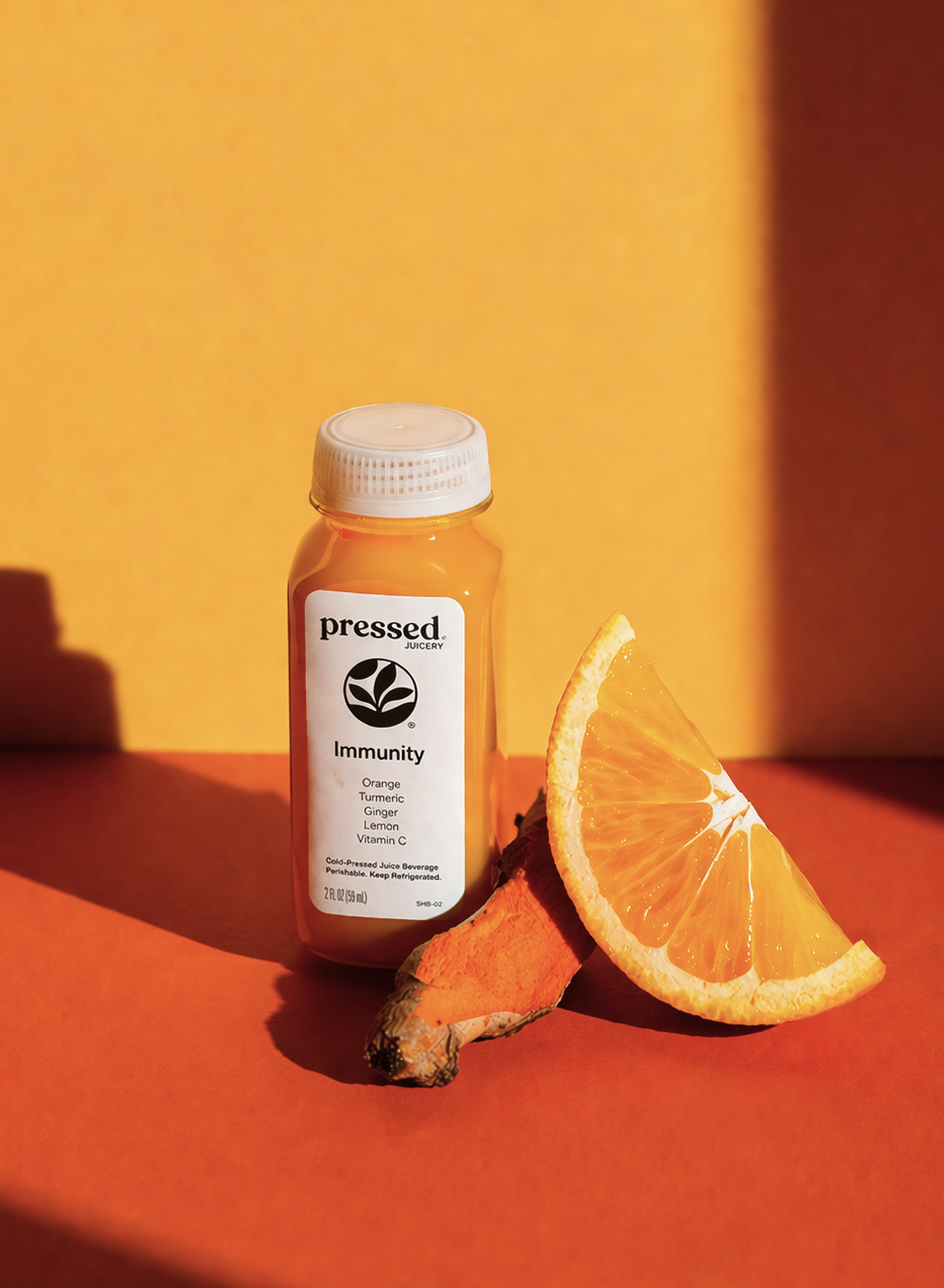 A bottle of Pressed Immunity Shot stands on a warm orange surface against a yellow-orange background, styled with a sliced orange and a root vegetable. The bottle label reads 'pressed Immunity Orange Turmeric Vitamin C'.